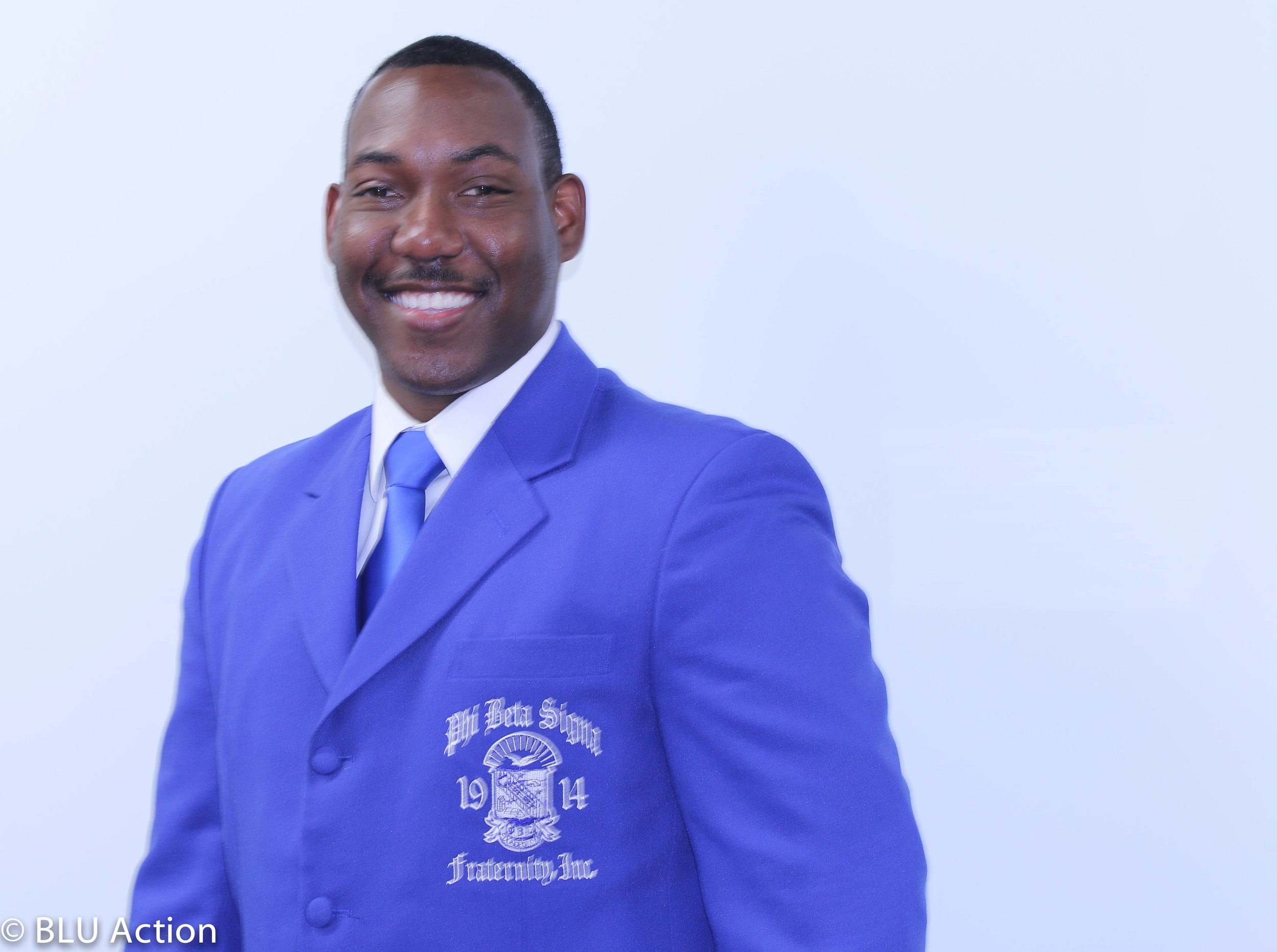 A smiling African American man in a blue blazer and tie posing against a white background.