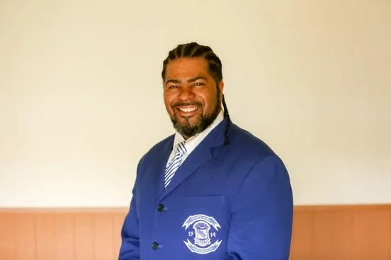 A man with dark hair and a beard wearing a blue blazer with a school or organization crest, smiling in front of a plain background.