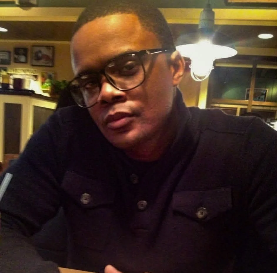 A young man wearing glasses and a black jacket sitting in a restaurant or cafe.