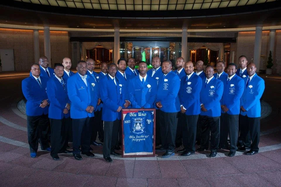Group of men dressed in blue blazers and black pants, standing together in a semi-circle, with a banner in front that reads "Blu Uno" and "Blue Inch of Nippon." They are in a well-lit, elegant venue with a curved ceiling and glass doors behind them.