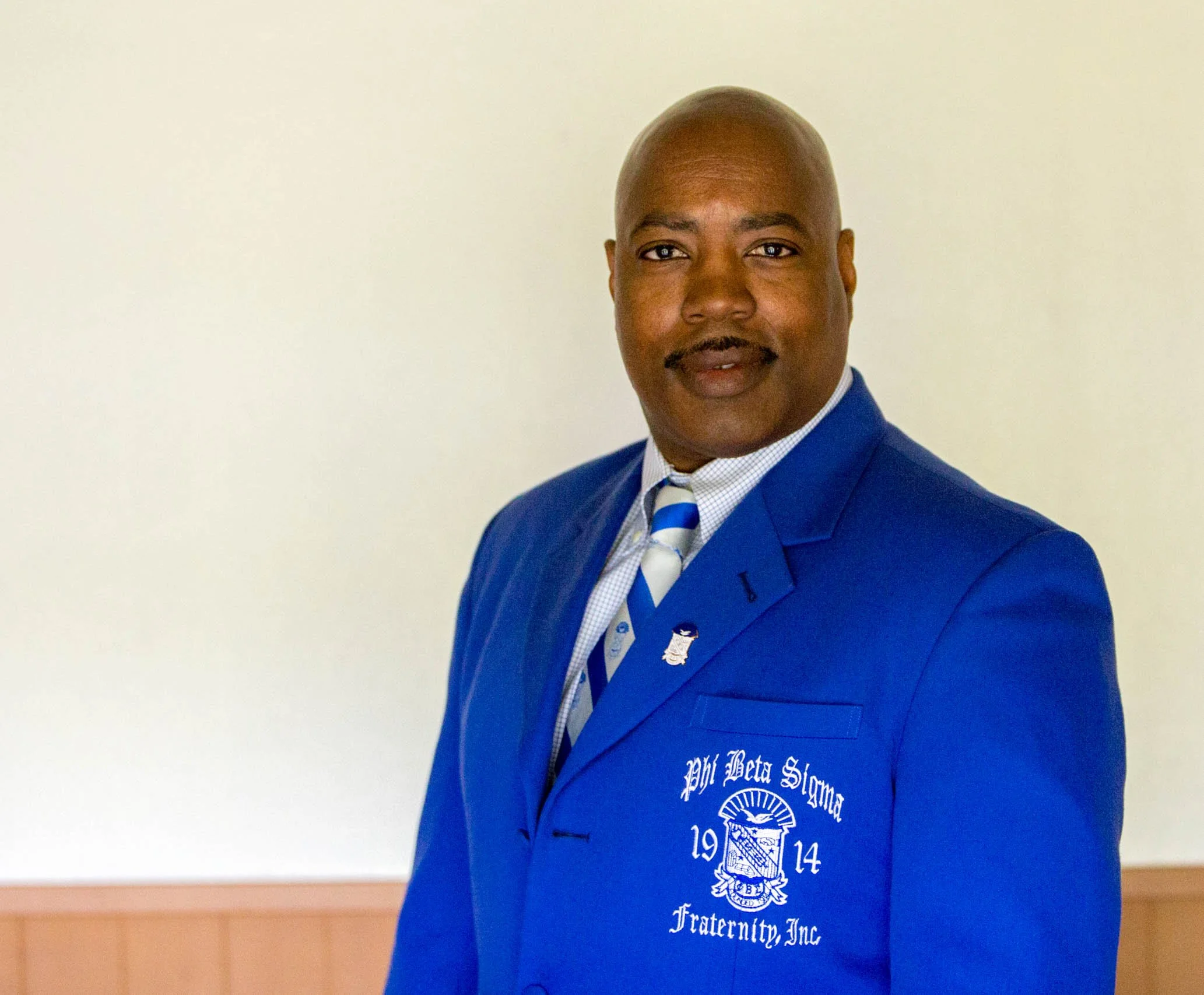 A man in a blue fraternity blazer with an embroidered crest and text, wearing a shirt and tie, standing against a plain background.
