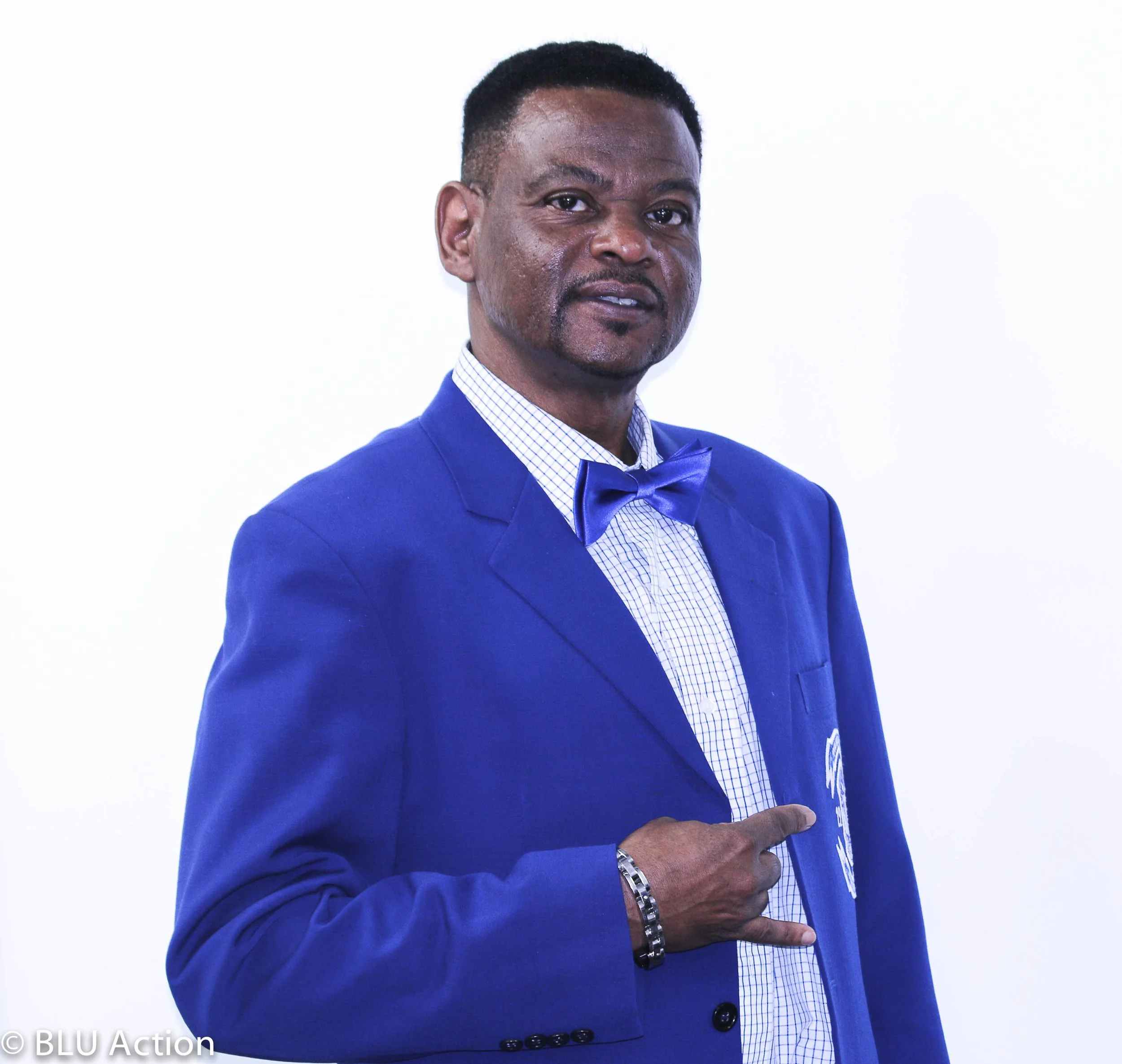 A man dressed in a blue suit with a matching bow tie, pointing at an emblem on his suit, standing against a plain white background.