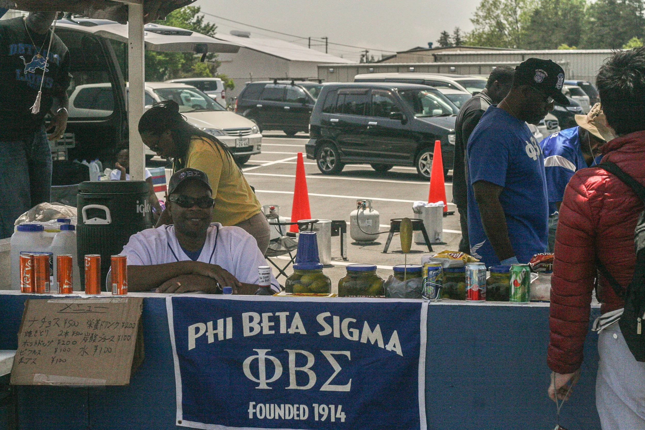 A group of people gathered around a table with drinks, snacks, and a banner that reads 'Phi Beta Sigma Founded 1914'. The scene is set in an outdoor parking lot with cars and orange traffic cones in the background.