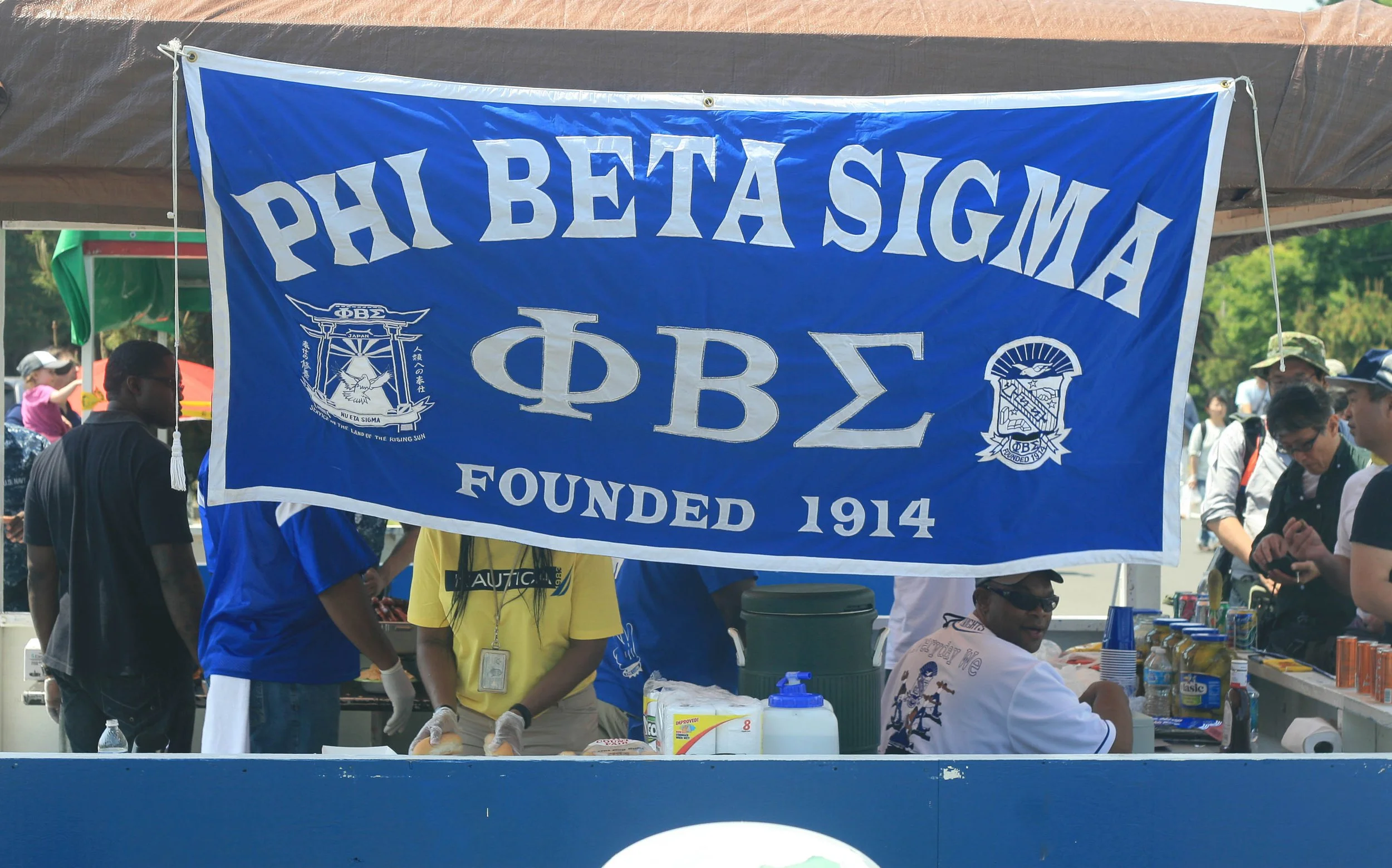 A group of people behind a table at a Phi Beta Sigma fraternity event, with a large blue banner displaying the fraternity's name, symbols, and founding year.