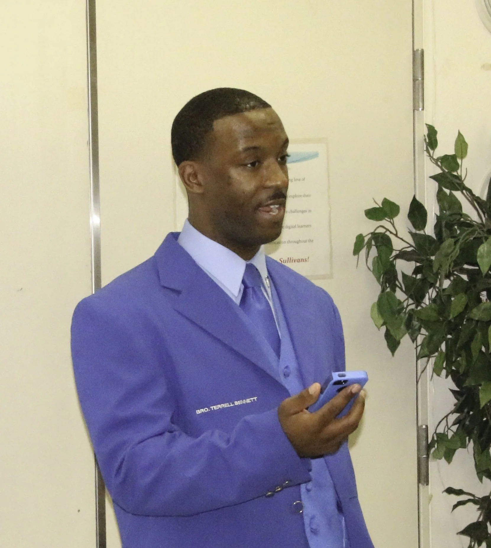 A young man dressed in a blue blazer and matching tie, holding a cellphone, standing indoors near a white wall and a green potted plant.