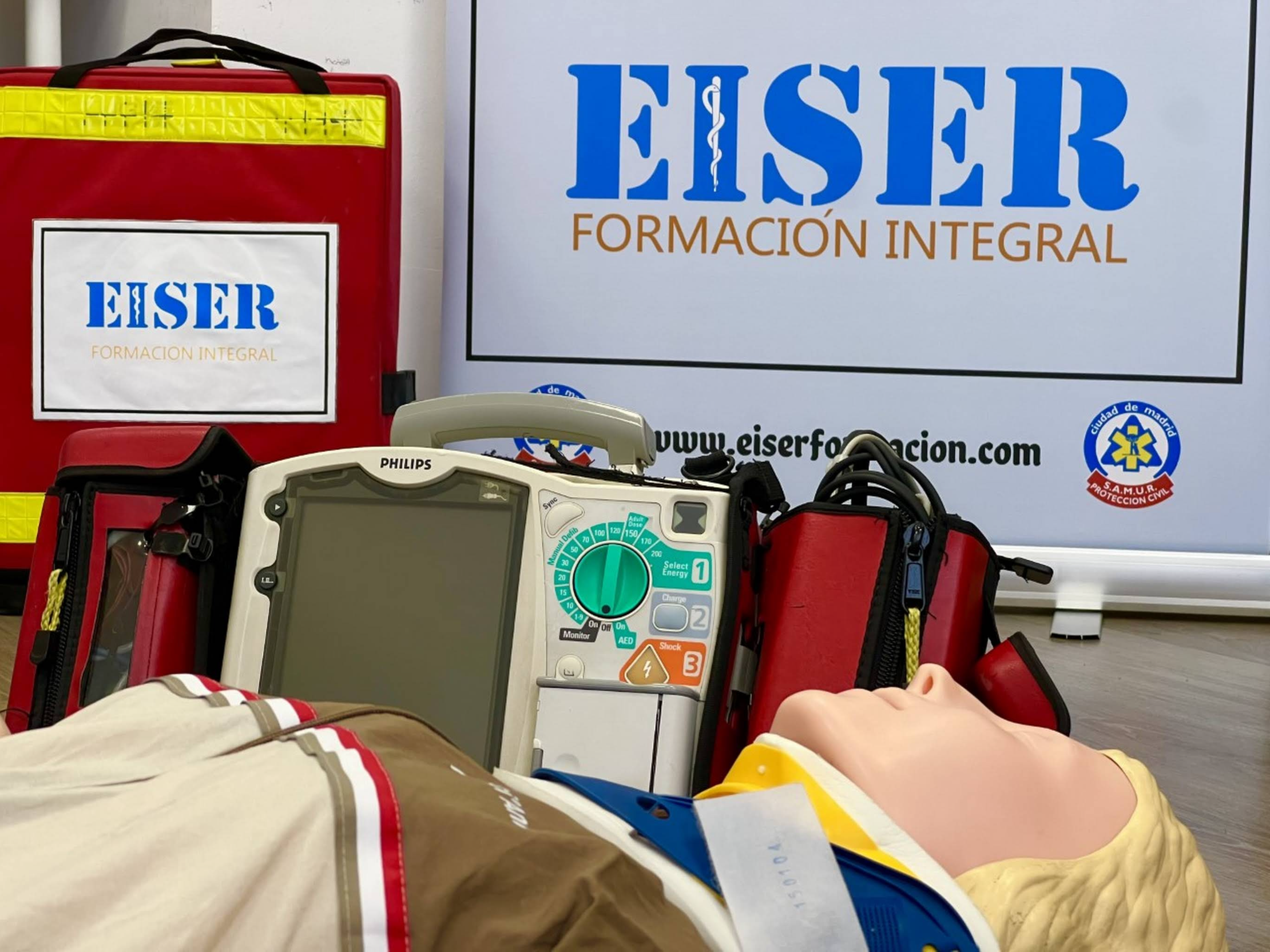 Equipo de primeros auxilios, incluyendo una máscara de reanimación, un monitor de signos vitales, una caja de emergencia roja y una cabeza de simulación para entrenamiento de rescate, en un entorno de capacitación con un cartel de EISER Formación Integral.