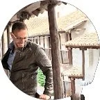 Hombre con gafas en una esquina de una estructura de madera, en un entorno exterior con techos de tejas.