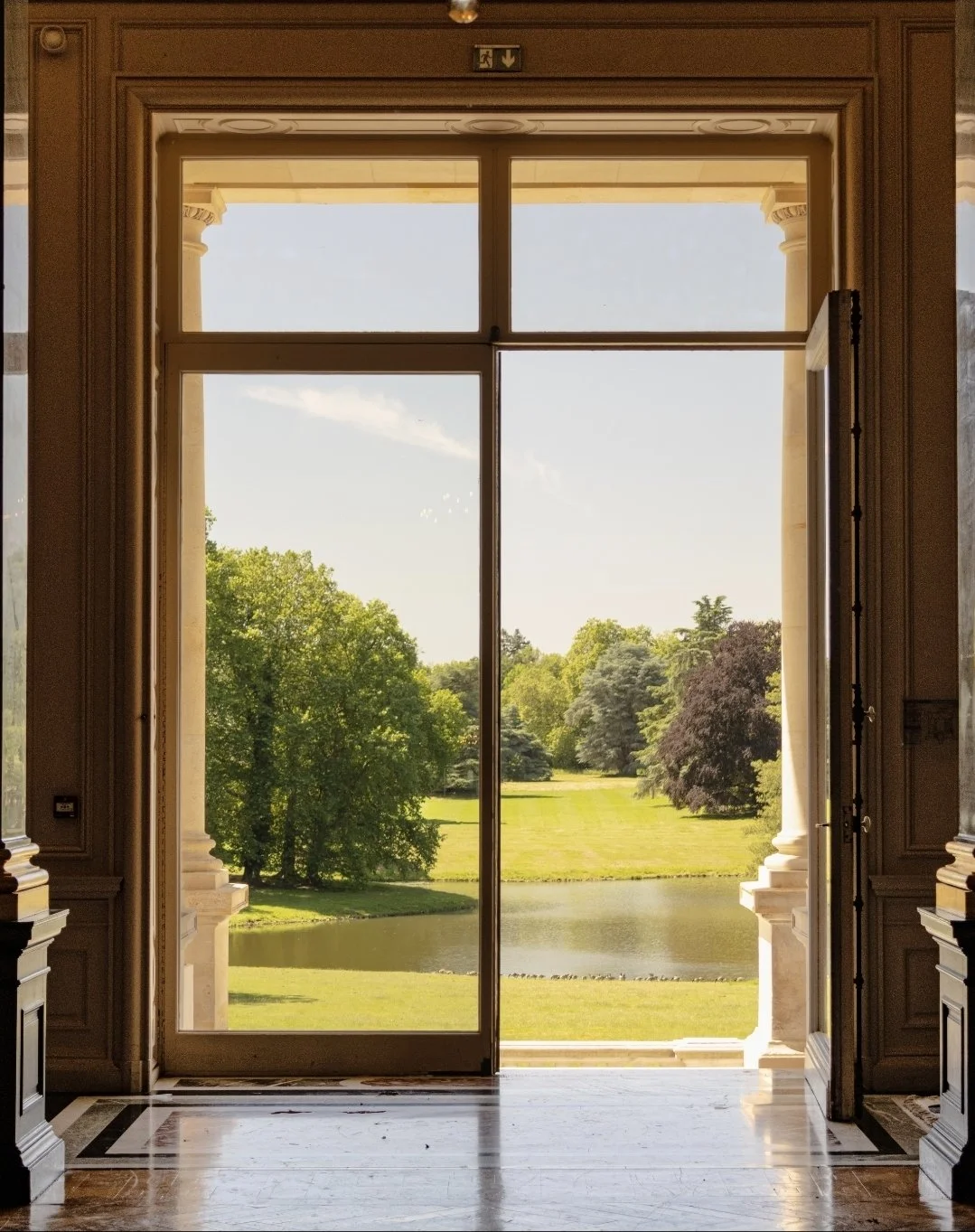 Vue d'une fenêtre grande donnant sur un jardin verdoyant avec des arbres et un étang, dans une salle lumineuse avec un sol en marbre.