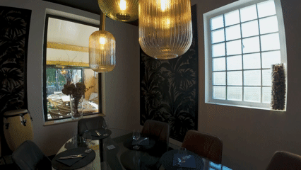 Salle à manger avec une table, des chaises, des suspensions lumineuses, un mur avec un motif de feuilles, et une fenêtre avec une grille.