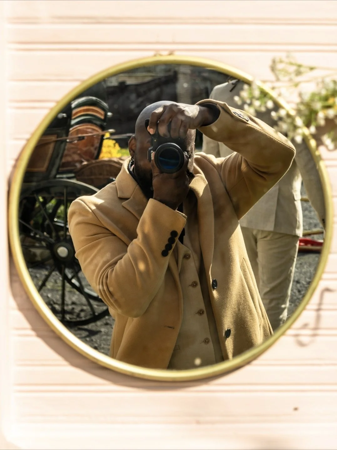 Homme prenant une photo avec un appareil photo devant un miroir rond, portant un manteau beige, avec un vélo et des fleurs en arrière-plan.
