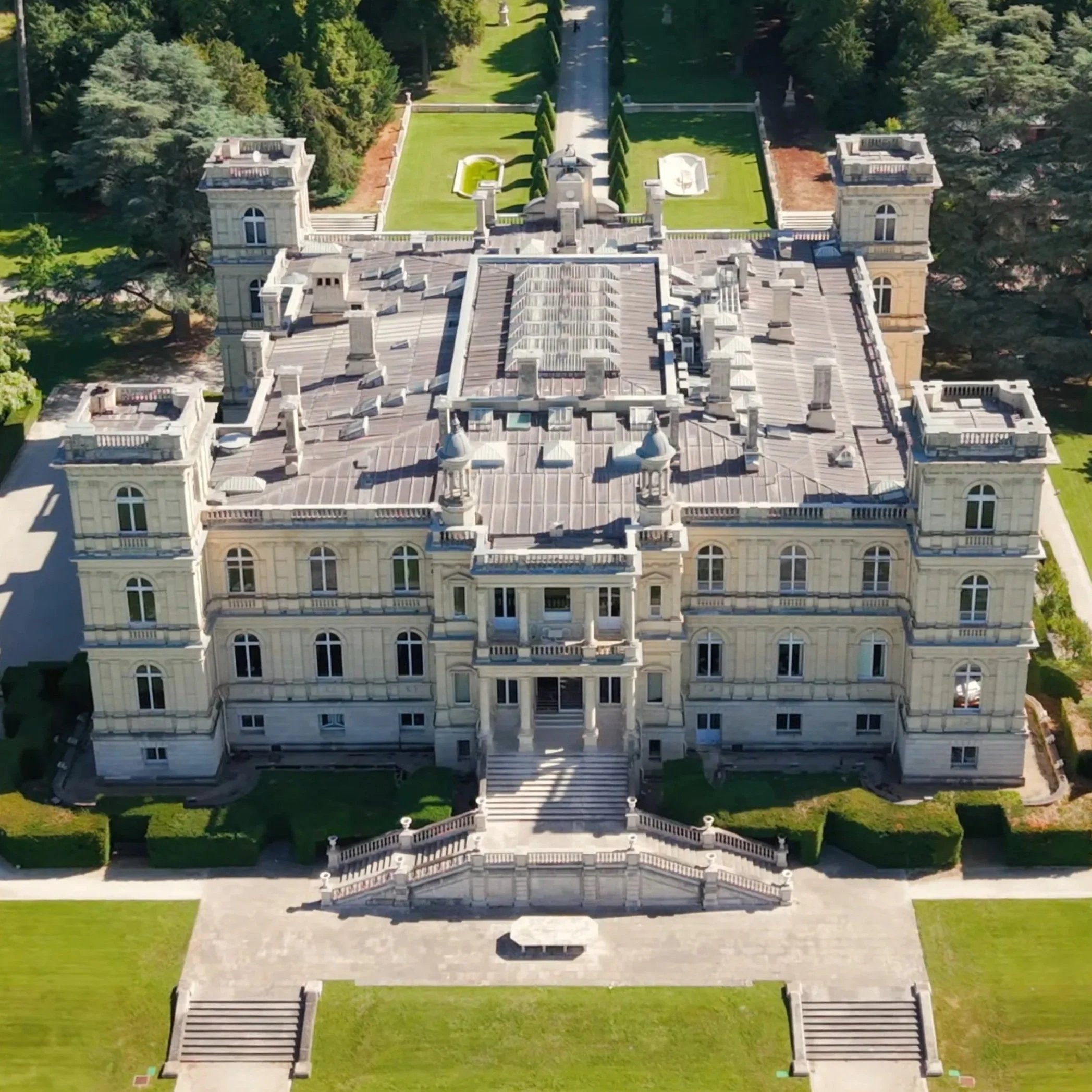 Vue aérienne d'un grand bâtiment de style classique avec plusieurs étages et une façade ornée, entouré de jardin bien entretenu avec des allées et des arbres.