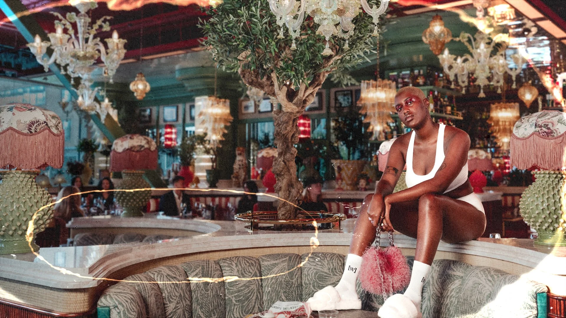 Stylish person in white outfit posing on a vintage sofa in a luxurious, ornately decorated restaurant with chandeliers and green decor.
