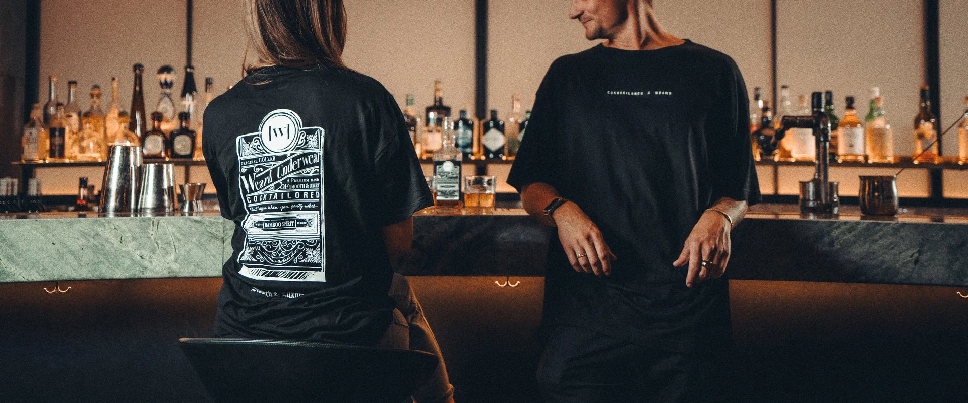 Two people wearing black T-shirts at a bar with various liquor bottles in the background. For the Weard Underwear x Cocktailored campaign.