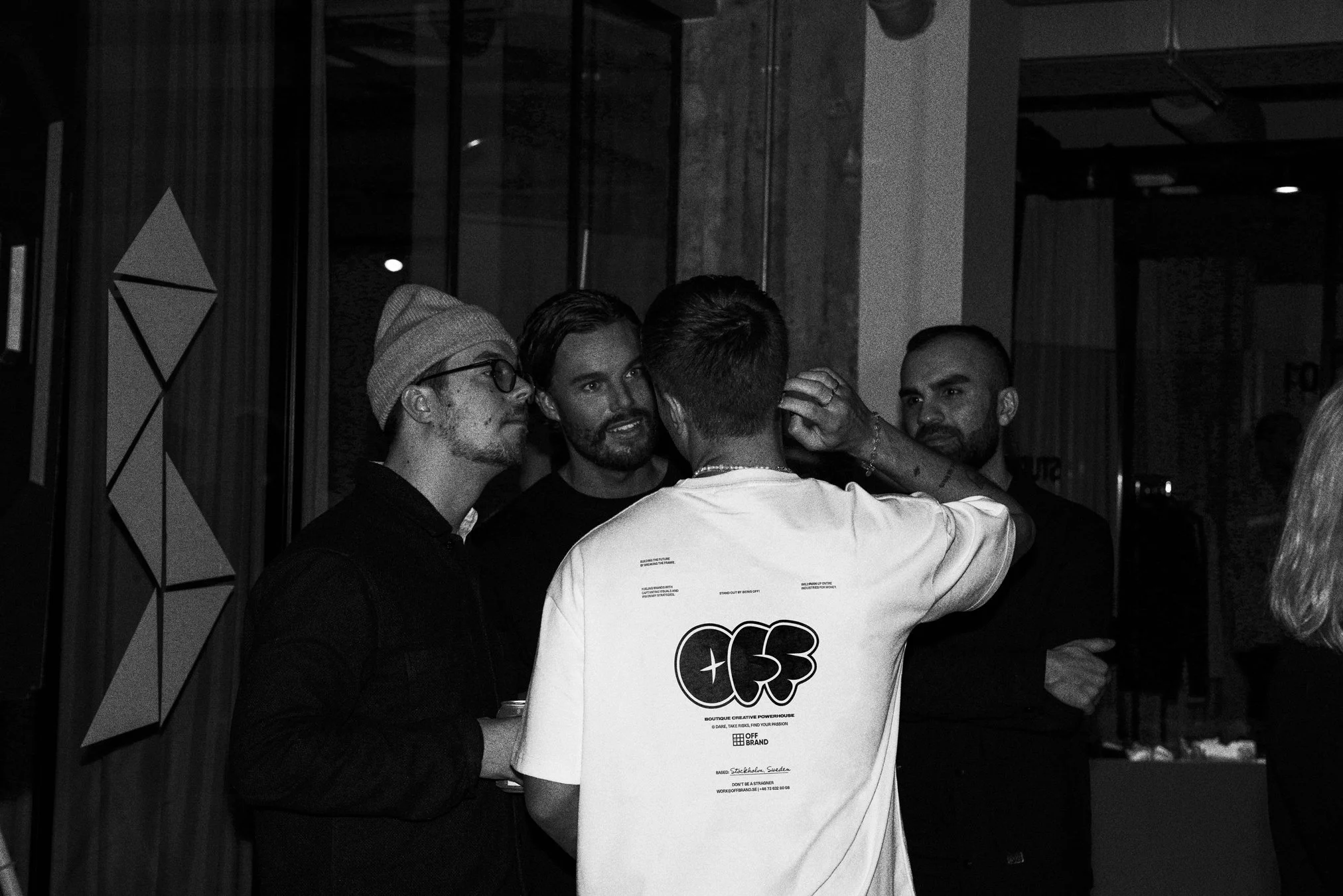 A group of four men engaged in conversation in a dimly lit indoor setting. One wears a beanie and glasses, another is facing away wearing a T-shirt with a graphic design on the back. The room has decorative panels and a modern interior design.