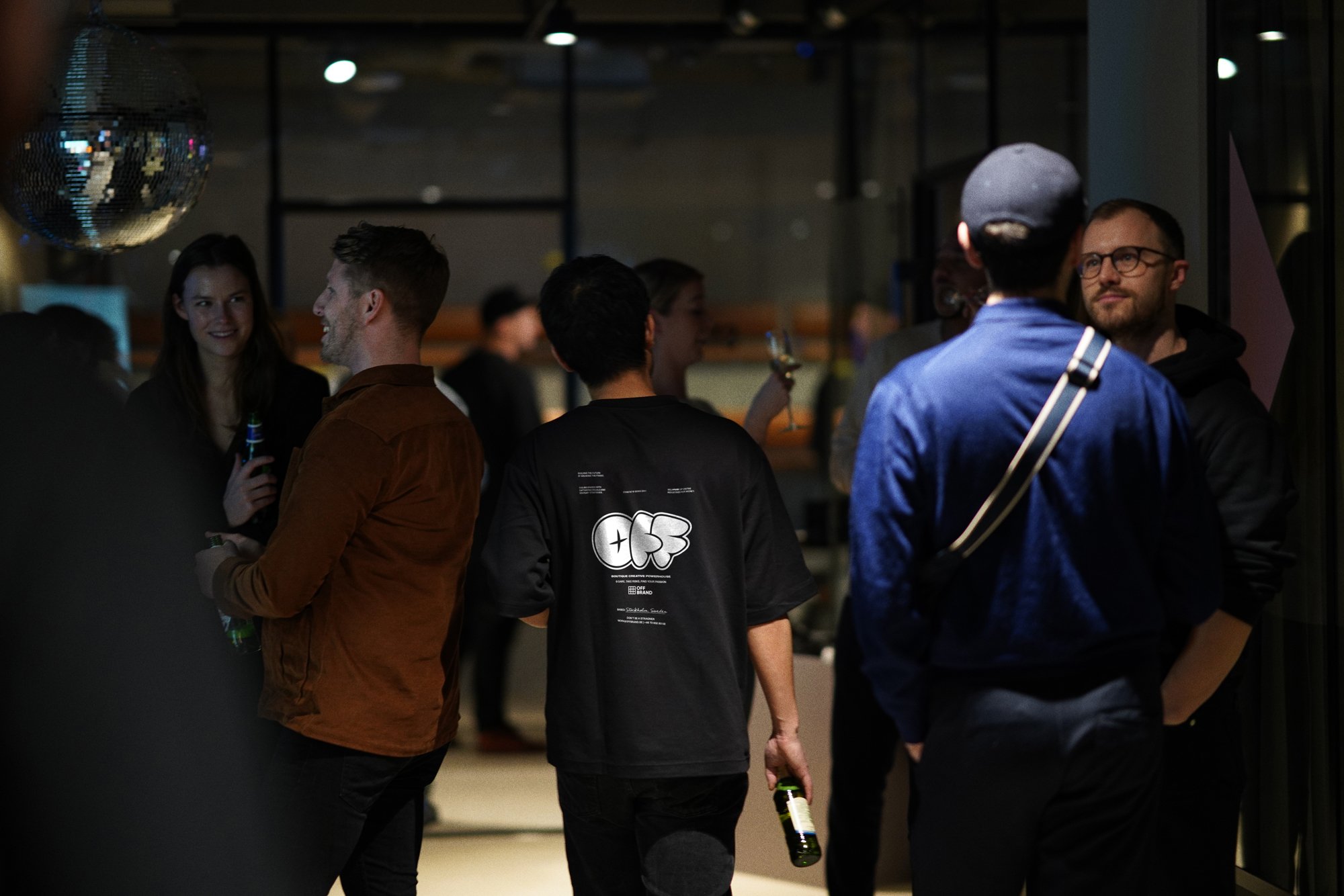Man in Off+brand t-shirt at a party. Group of people socializing in a dimly lit indoor space, some holding drinks.