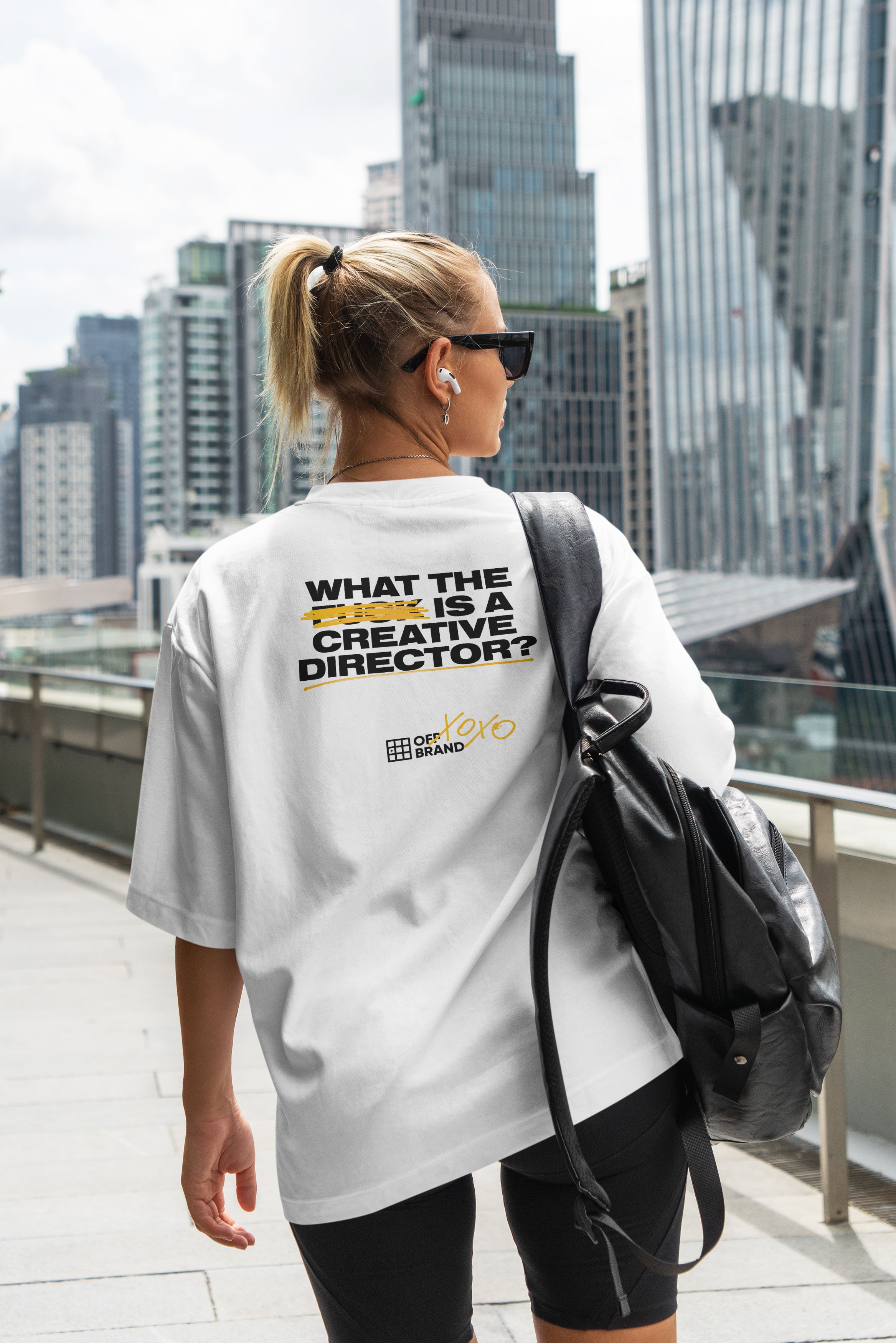 Person walking on a city walkway, wearing a white T-shirt with text "What the [censored] is a creative director?" from OFF+BRAND. Carrying a black backpack and wearing sunglasses and earbuds.