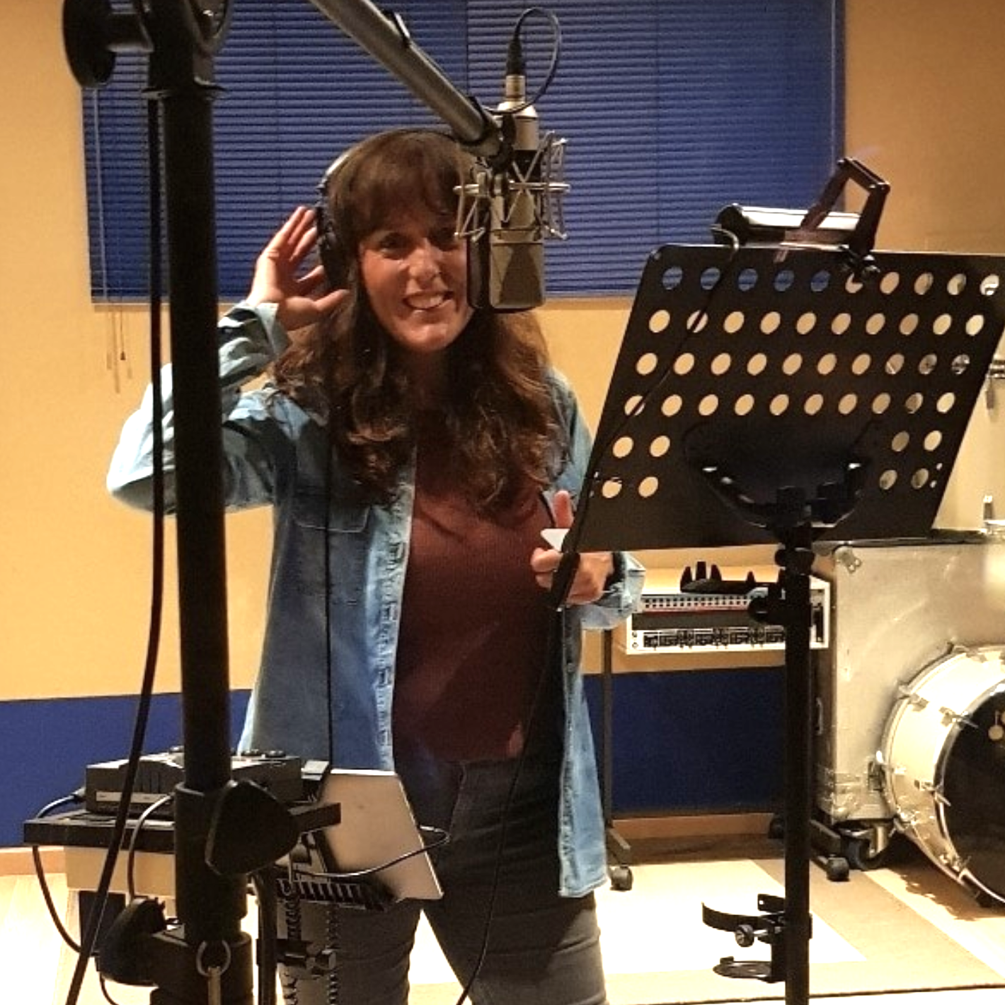 Woman in a recording studio with headphones, standing in front of a microphone, holding a script, and smiling.