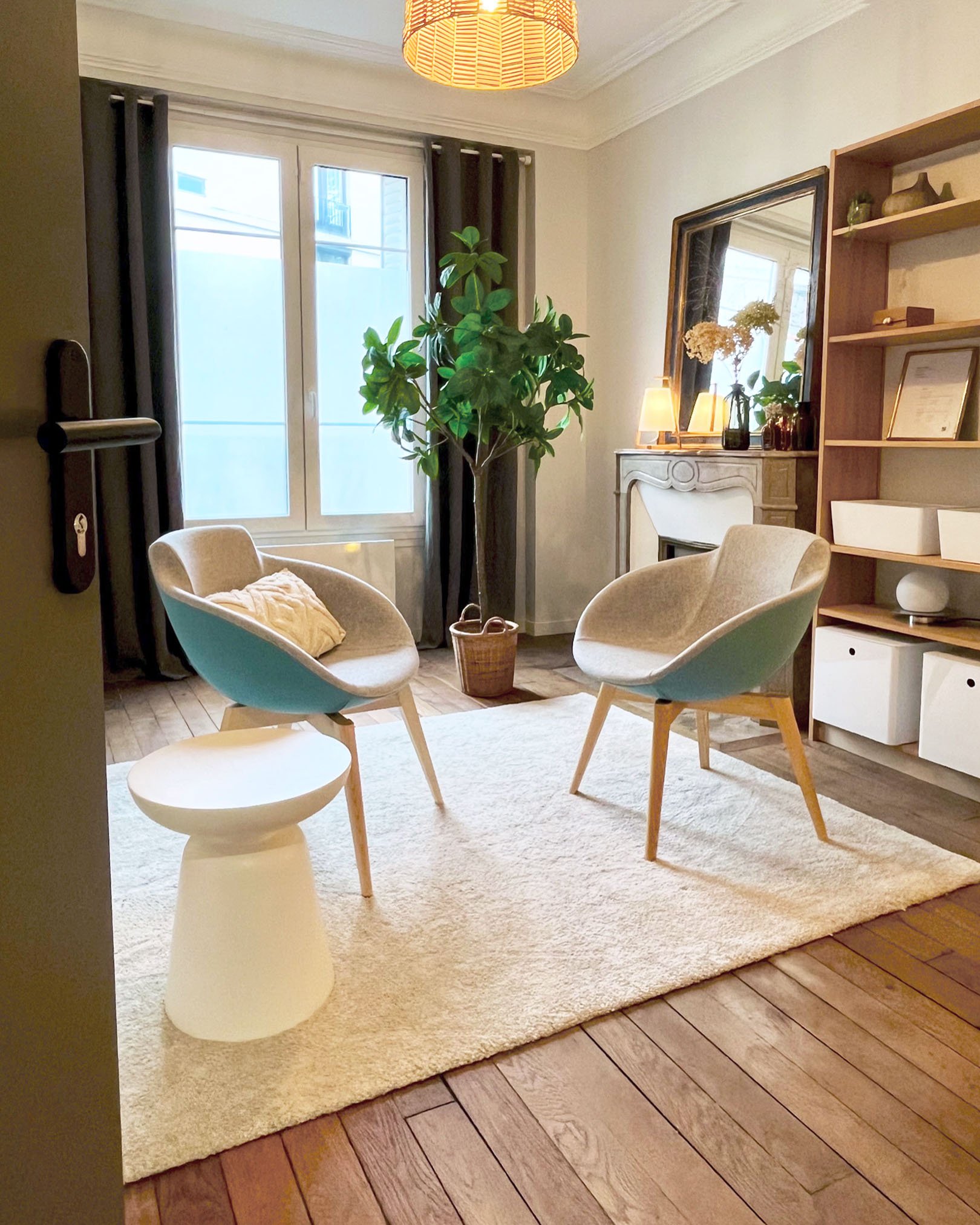 Salon avec deux fauteuils, une plante d'intérieur, un tapis beige, une cheminée, un miroir, une étagère en bois, et une petite table blanche.