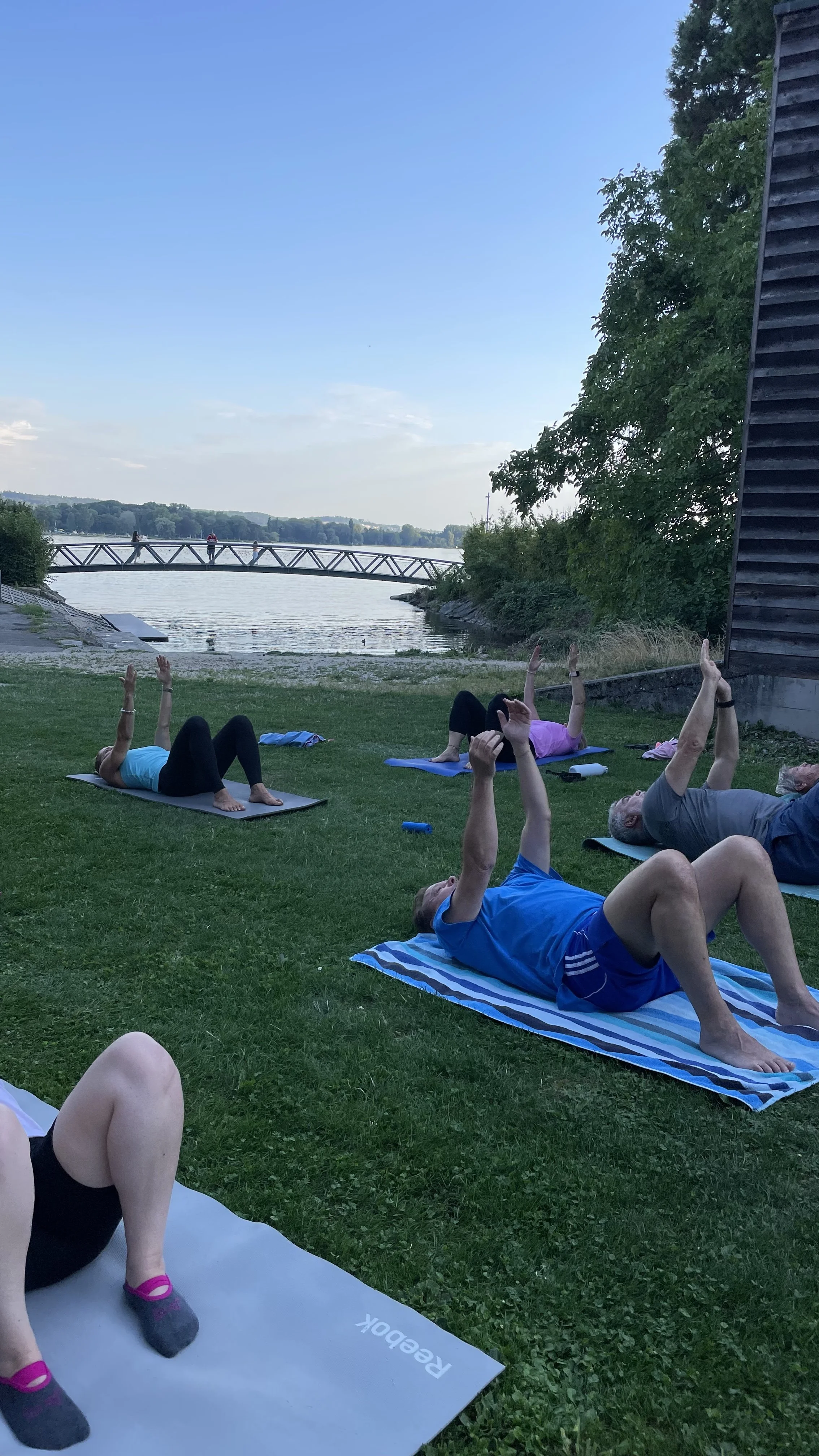 Menschen machen Yoga im Freien auf einer Wiese am Fluss bei Sonnenuntergang, umgeben von Bäumen, mit einem Fußgängerbrücke im Hintergrund.