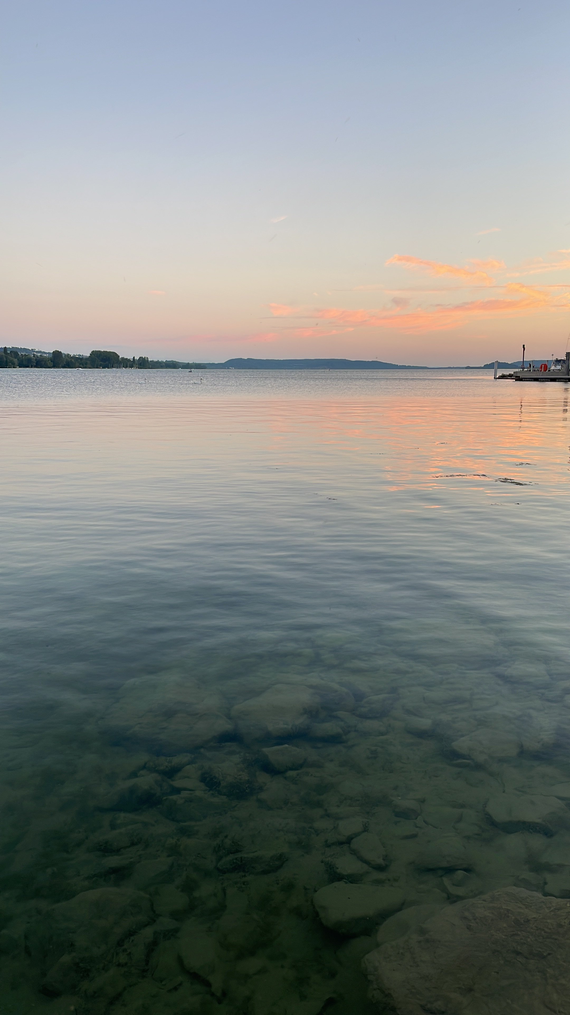 Ein ruhiges Wasser mit sichtbaren Steinen im Flachwasserbereich, ein klares Gewässer mit spiegelnder Oberfläche, im Hintergrund ein Himmel bei Sonnenuntergang mit leicht rosa und orangefarbenen Wolken, ein Ufer mit Bäumen und ein Boot am rechten Ufer.