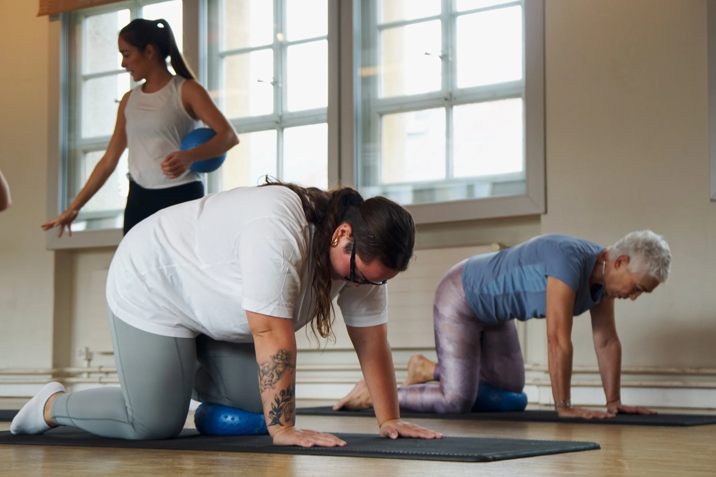 Menschen bei einer Yoga- oder Pilates-Übung auf einer Matte im Raum, bei der die Personen in einem Vierfüßlerstand sind, während eine Frau mit einem blauen Ball neben ihnen steht.