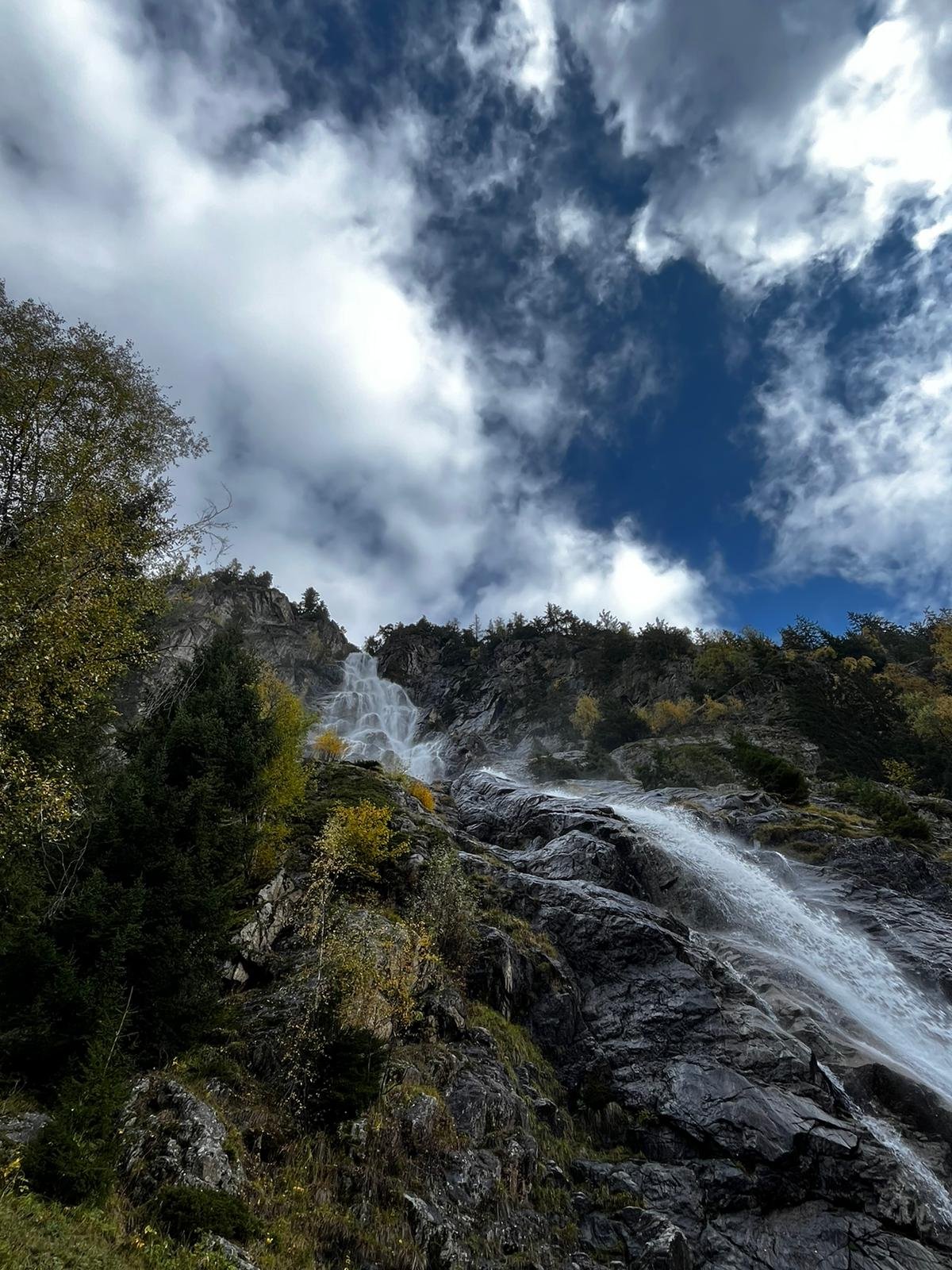 Ein Wasserfall fließt die felsige Bergwand hinunter, umgeben von Bäumen, unter einem teilweise bewölkten Himmel.