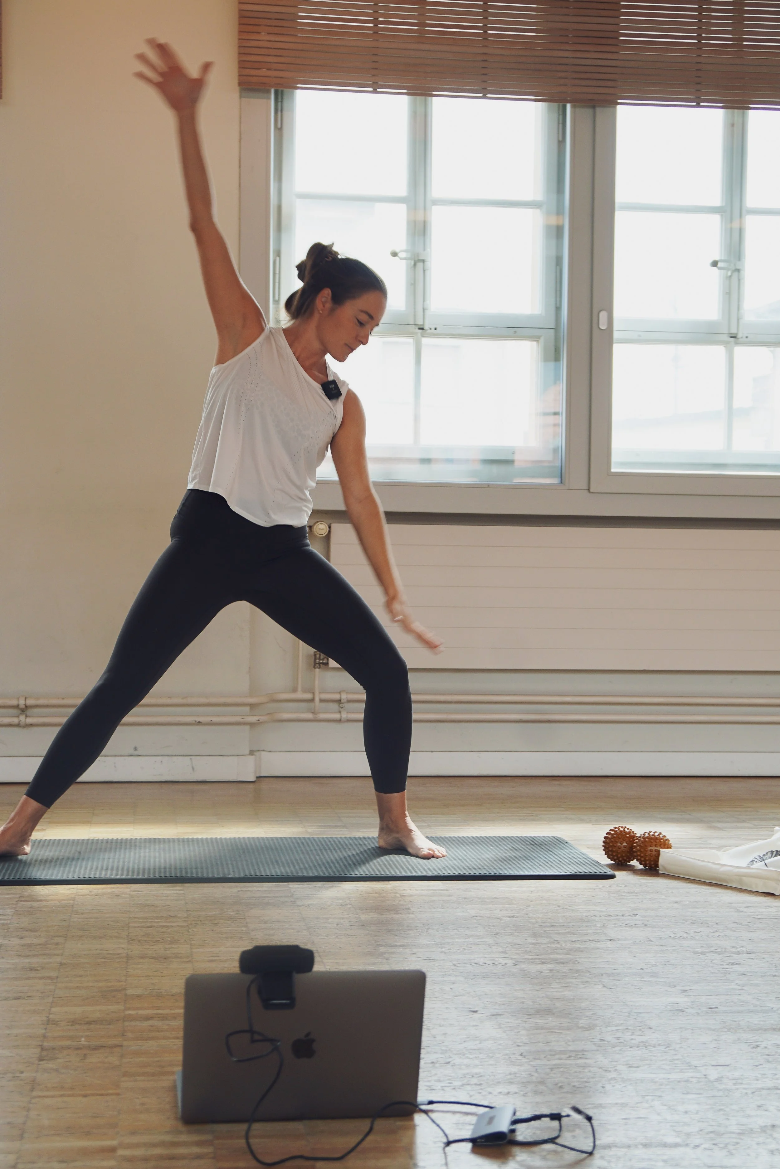 Eine Frau macht Yoga im Wohnzimmer vor einer großen Fensterfront, trägt Sportkleidung, steht in der Warrior-I-Position und bewegt einen Arm nach oben. Auf dem Boden liegen Rutschmatte, kleine Massagebälle und eine Rolle. Ein Laptop ist vor ihr, vermutlich für eine Online-Workout- oder Yoga-Stunde.