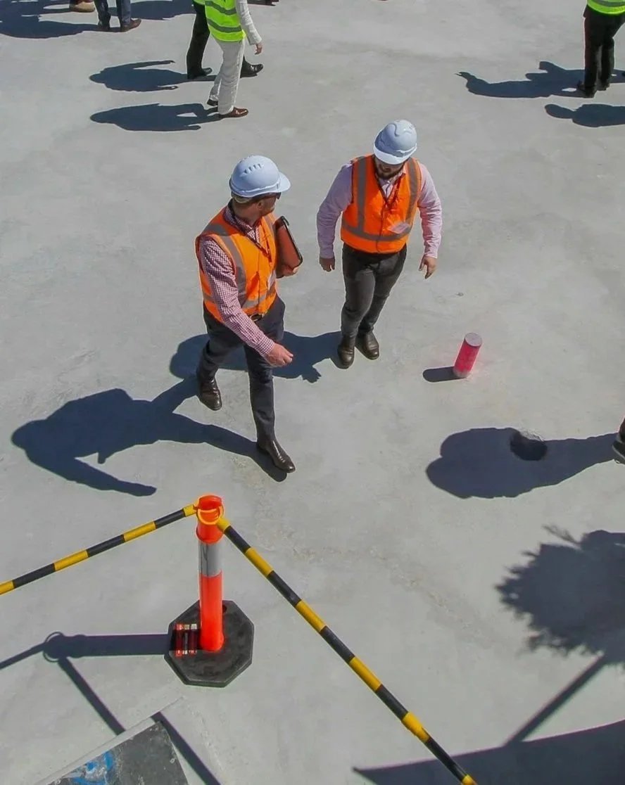 Building Inspections. Two construction workers wearing safety vests and hard hats standing on a construction site, with construction cones and barrier tape visible.