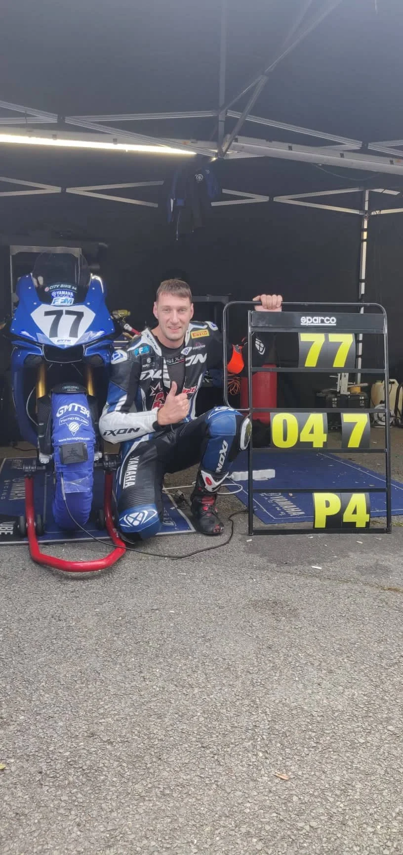 Un homme en combinaison de course de moto, assis près d'une moto Yamaha bleu avec le numéro 77, sous un stand de pit avec une roue à outils portant le même numéro.