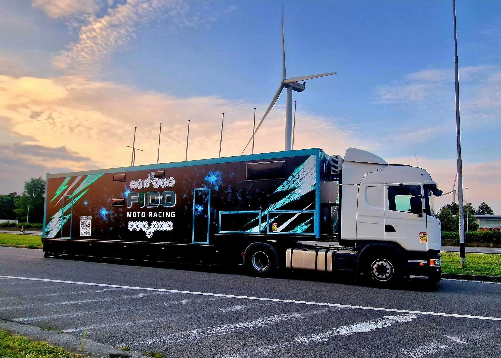 Camion de course moto avec une cabine blanche, décoré avec des motifs noirs et bleus, logo FIDO Moto Racing, éolienne en arrière-plan, ciel au crépuscule.