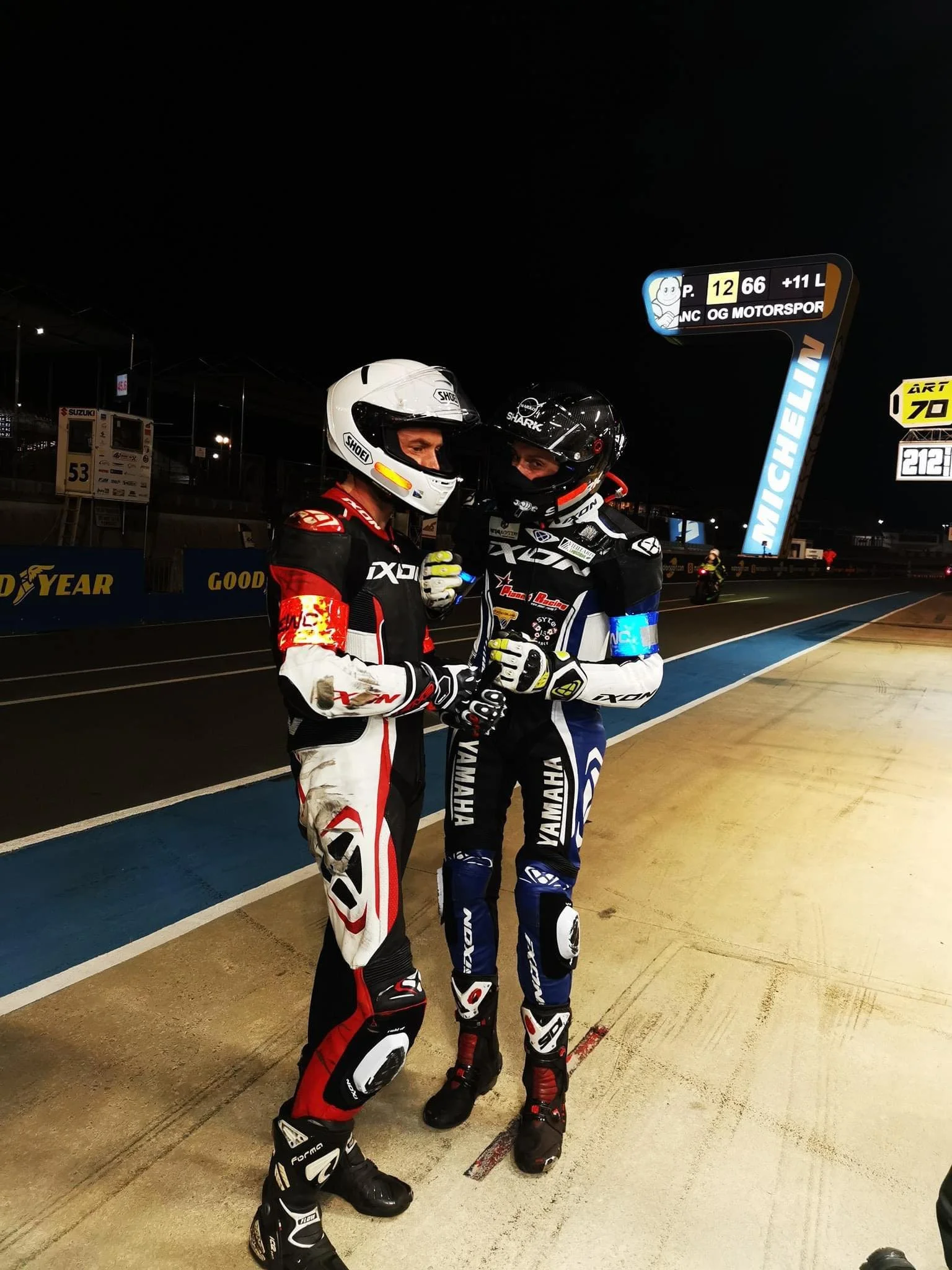 Deux pilotes de course de motos en tenue de course se serrent la main dans une zone de paddock de nuit, devant un panneau d'informations du circuit Michelin, avec d'autres personnes en arrière-plan.