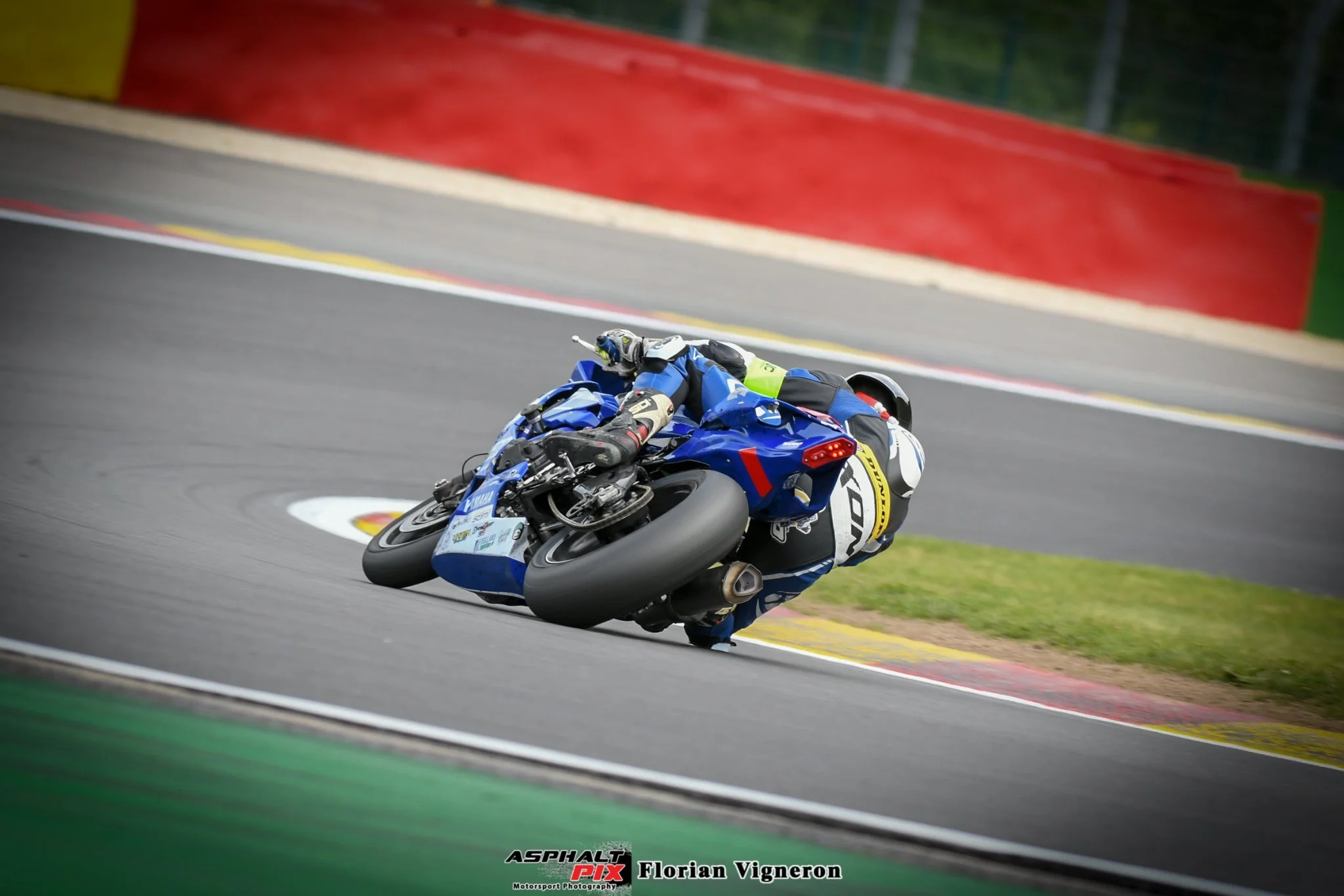 Un pilote de moto en pleine courbe lors d'une course sur un circuit asphalté, portant un équipement de protection bleue et un casque, avec un fond de barrières de course rouges et vertes.
