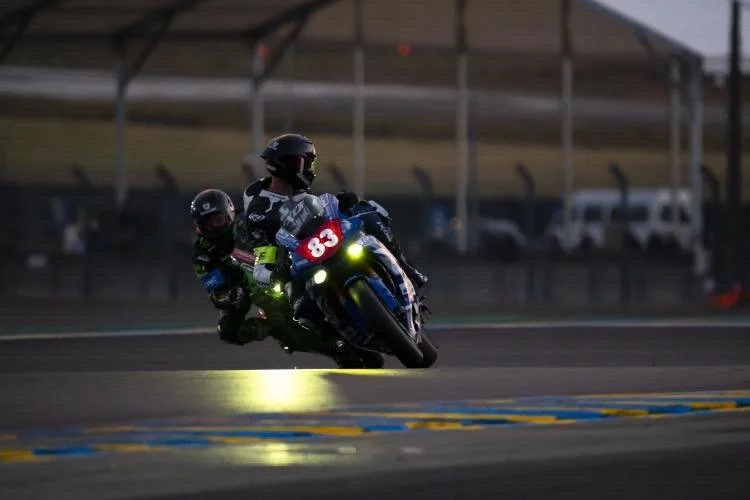 Deux motards en course, l'un avec le numéro 83, roulant à grande vitesse sur une piste de course.