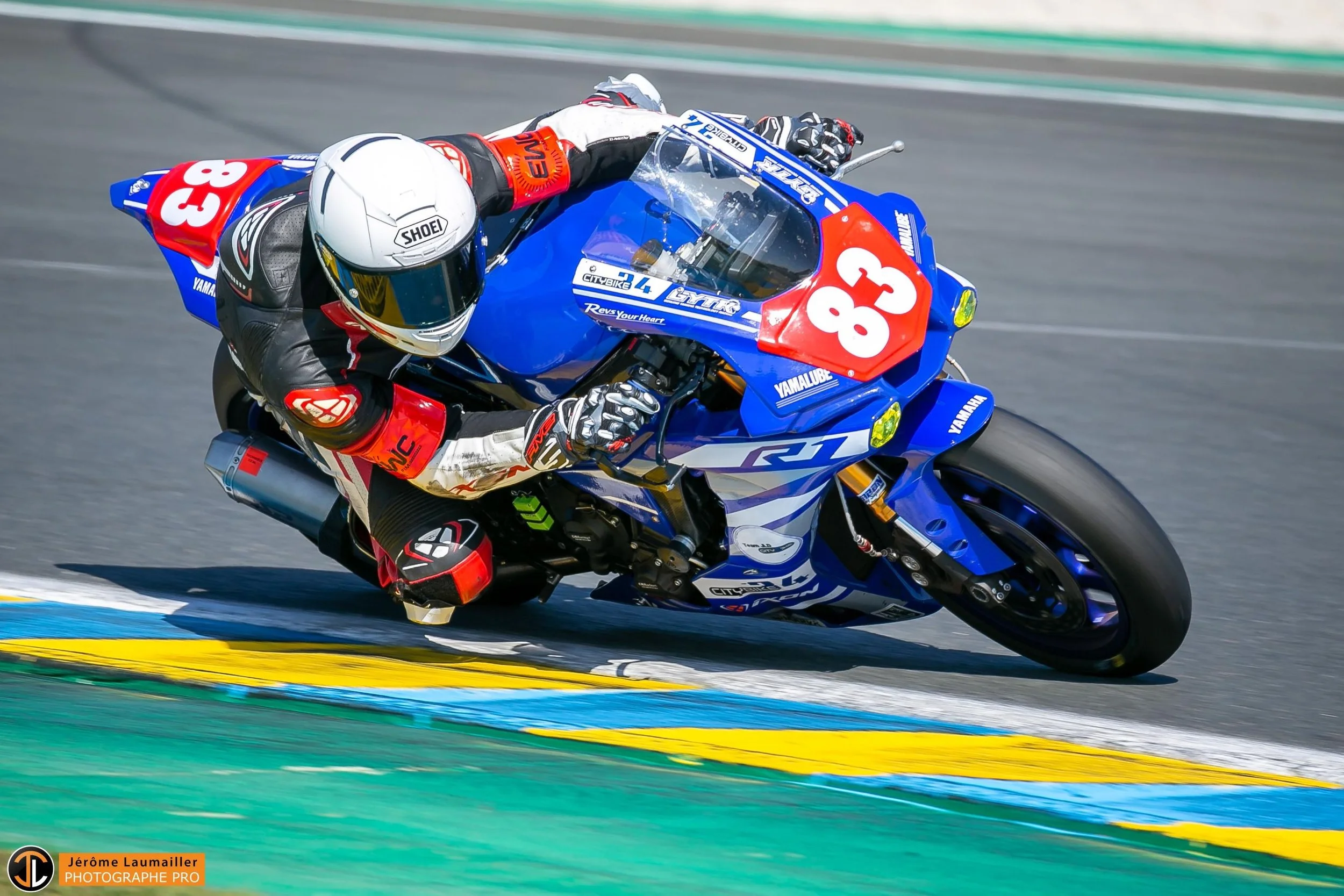 Un pilote de course en moto Yamaha R1, portant un casque blanc, bien incliné lors d'une course sur circuit, avec le numéro 83 sur la moto.