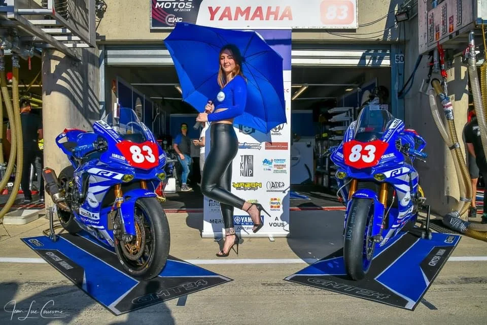 Une femme souriante pose devant deux motos de course Yamaha, portant un parapluie bleu et une tenue élégante, dans une zone de stands de motos de course.