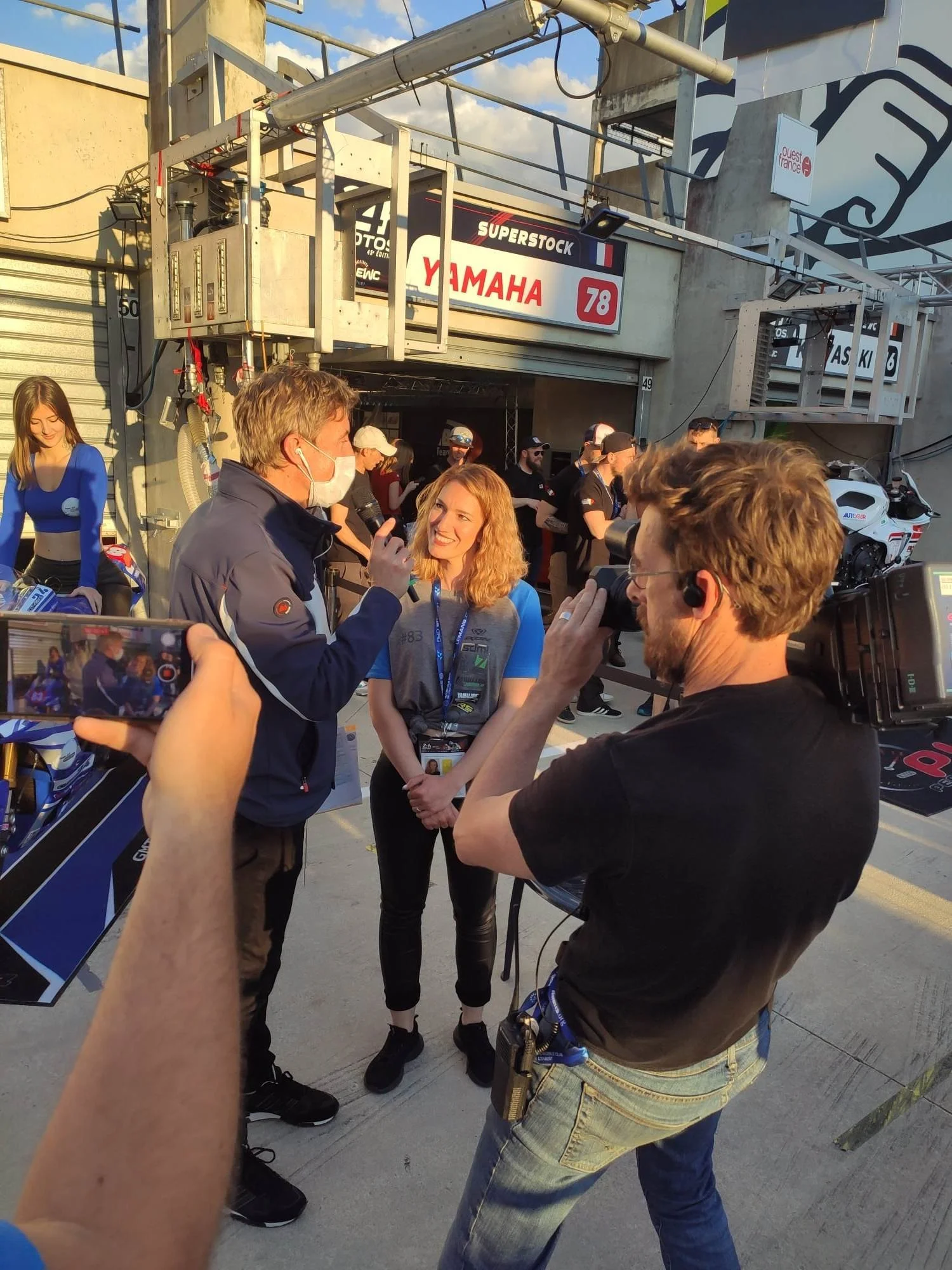 Interview entre une femme et un homme portant un masque face à une caméra, avec une personne prenant une photo ou vidéo, entourés d'autres personnes dans un environnement de garage ou de paddock, avec des affichages de marques de motos.