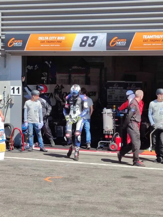 Un pilote de moto portant une combinaison de course et un casque, se dirigeant vers la piste de course dans un paddock, entouré d'autres membres d'équipe et de techniciens, devant un garage de Moto à l'occasion d'une compétition.