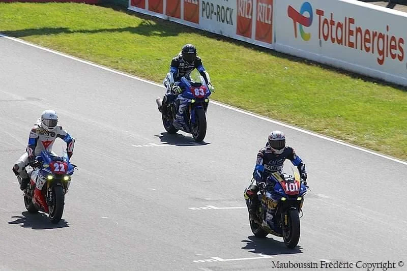Trois motards en course sur une piste, portant des combinaisons de course et casques, avec une barrièrepublicitaire en arrière-plan.