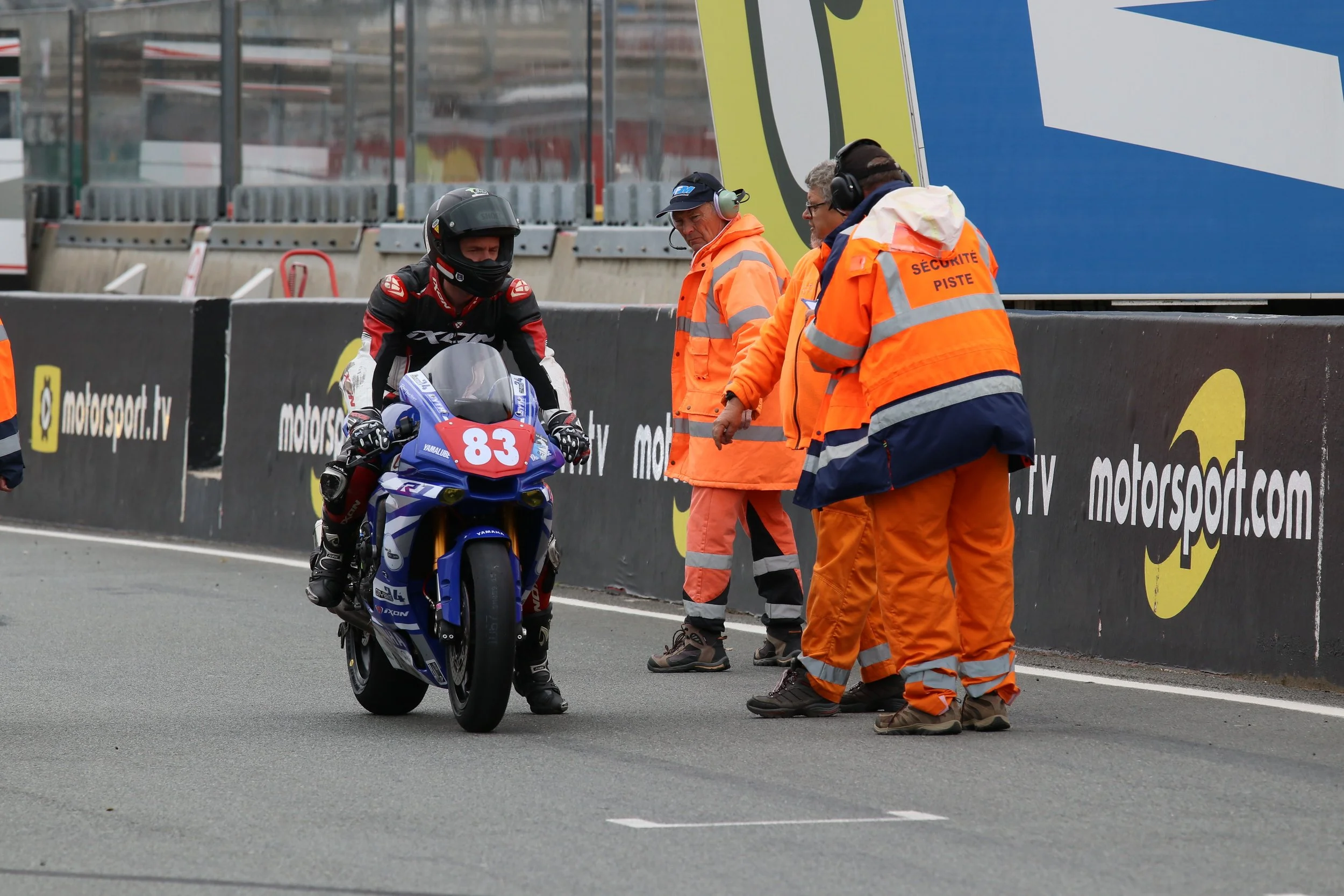 Un pilote de moto en tenue de course noire et rouge, portant un casque, positionné sur une moto bleue et blanche avec le numéro 83, est contrôlé par plusieurs hommes en blousons orange de sécurité sur une piste de course.