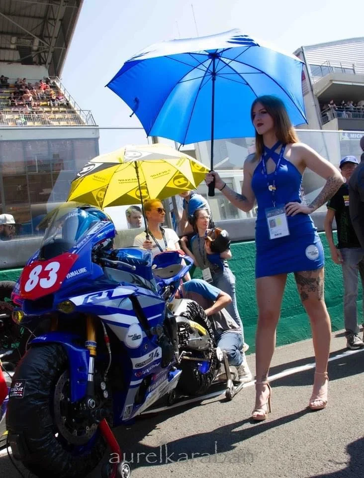 Une femme en robe bleue, portant un badge. Elle tient un parapluie bleu. Il y a une moto de course bleue avec le numéro 83. La scène se déroule dans un circuit avec plusieurs personnes en arrière-plan.