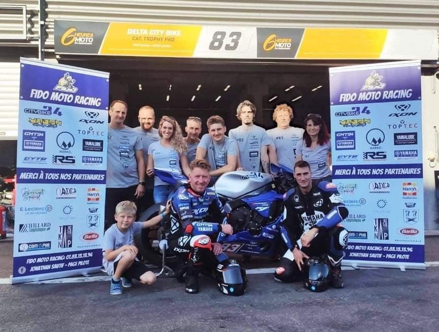 Groupe de personnes autour d'une moto de course Yamaha, avec deux pilotes en combinaison de course, lors d'un événement de moto racing.