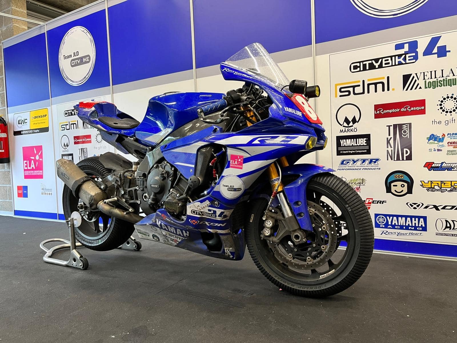 Une moto de course Yamaha bleue et blanche exposée dans un stand de vitrine, avec divers logos et publicités en arrière-plan.
