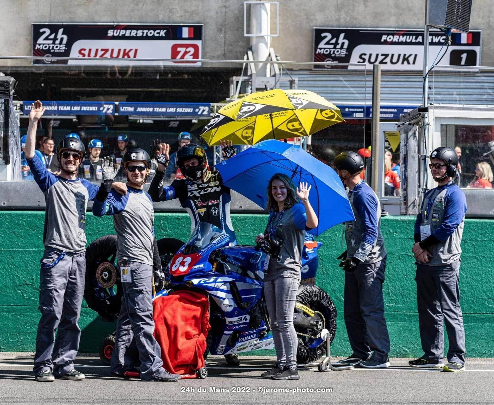Équipe de course moto posant dans la zone de pit, avec une moto bleue, des membres portant des casques et des vestes, certains tenant des parapluies, lors de l'événement 24h du Mans 2022.