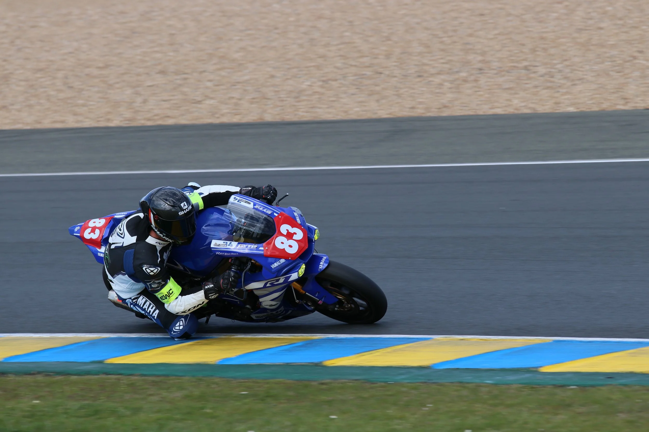 Un pilote de moto en course inclinée dans un virage serré sur une piste asphaltée, portant un casque noir et une combinaison de course, sur une moto Yamaha bleu avec le numéro 83.