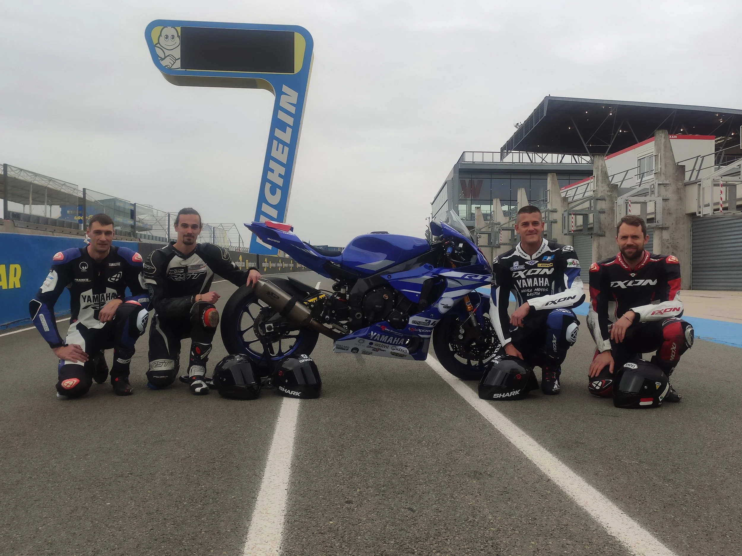 Cinq motos ambitionnées Ferrari et quatre pilotes en tenue de course sur une piste de course, posant devant une moto bleue de course Yamaha, avec un bâtiment et un panneau de sport automobile en arrière-plan.