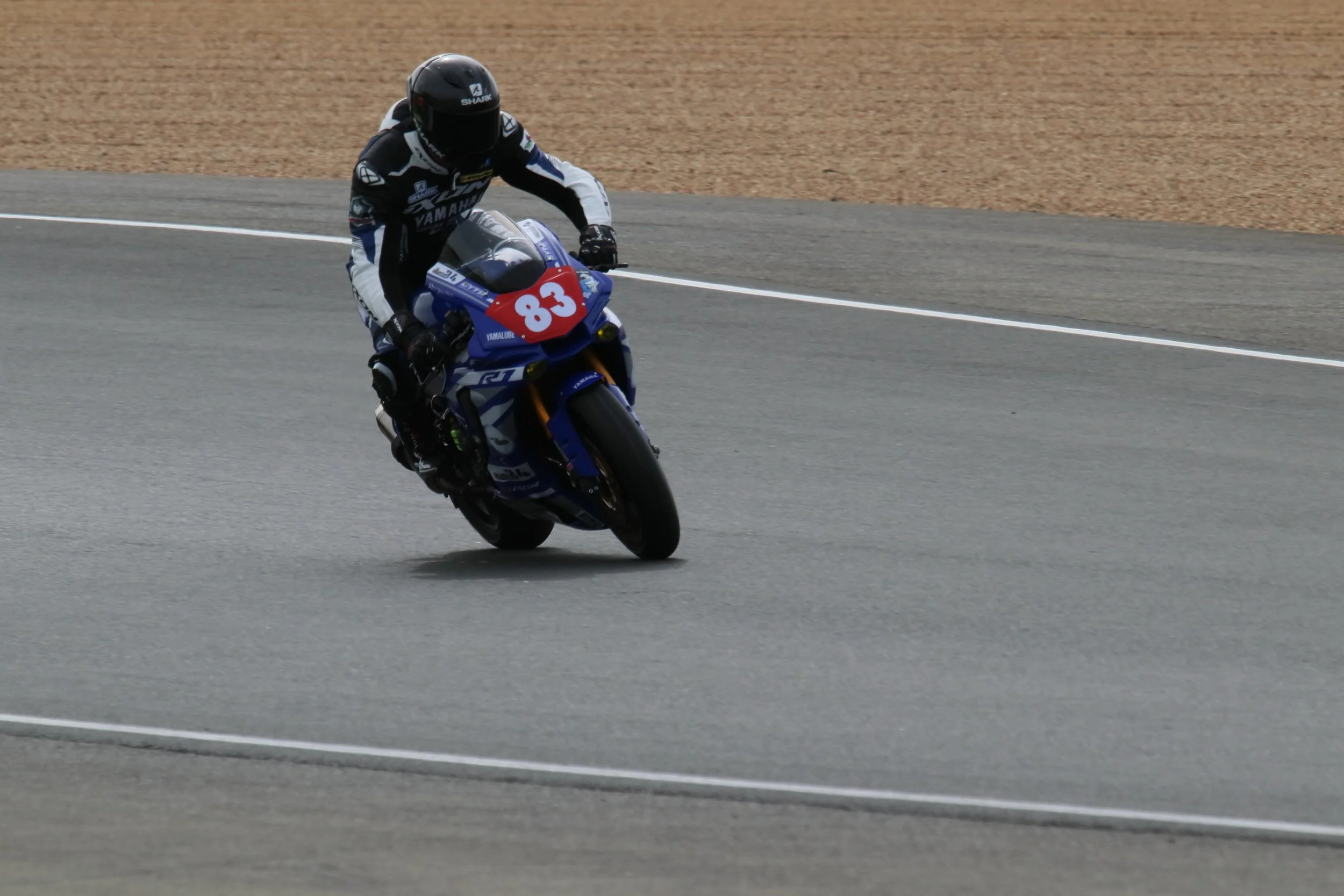 Un pilote de moto portant un casque noir et un équipement noir et blanc, en train de prendre un virage à grande vitesse sur une piste de course, monté sur une moto bleue avec le numéro 83 en rouge.