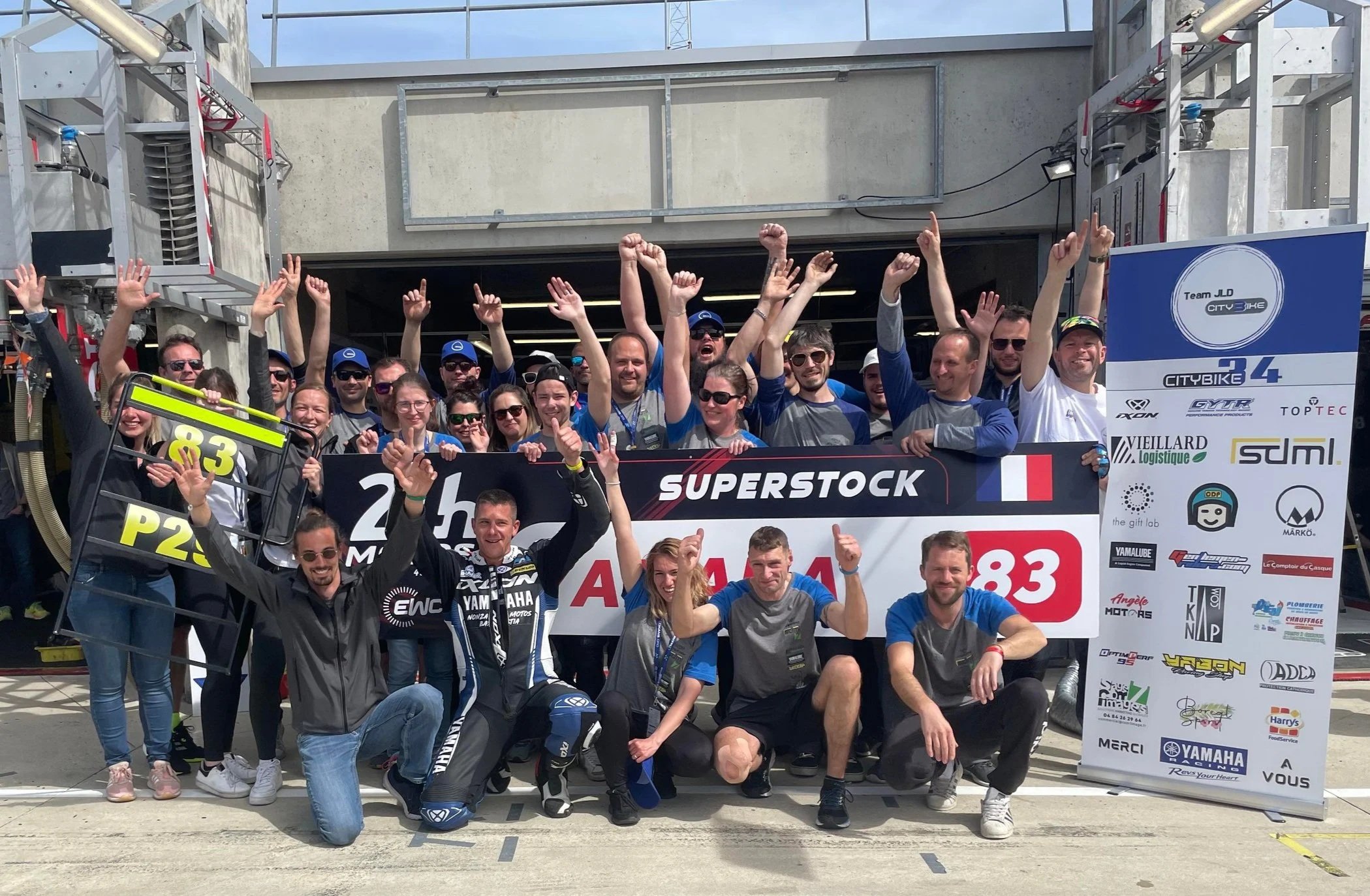 Groupe de personnes celebrant une victoire en équipe l'arrivée des 24hrs du Mans moto en face d'un panneau de sponsors.