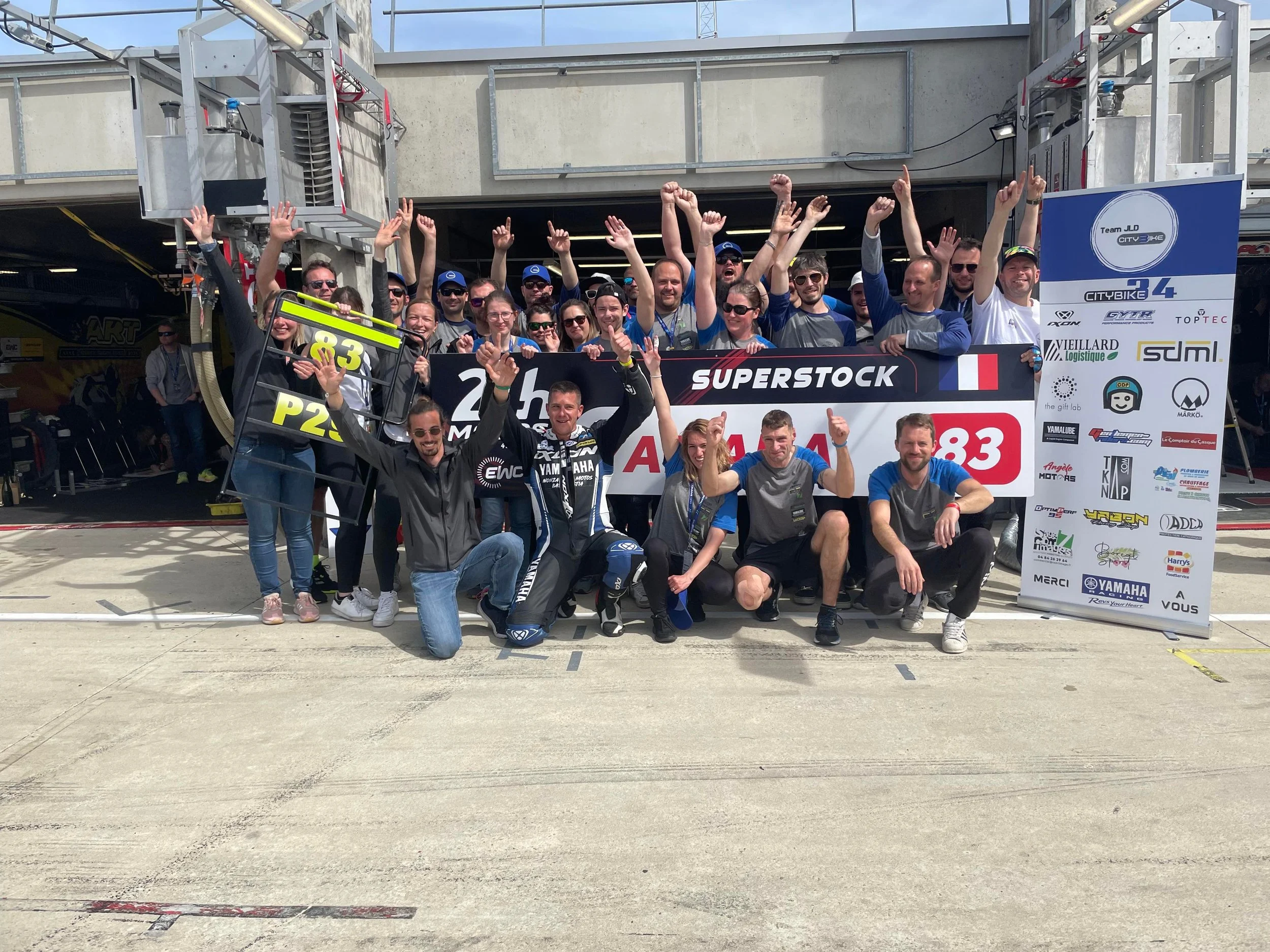 Groupe de personnes posant avec des panneaux dans le paddock d'une course de motos, célébrant la réussite.