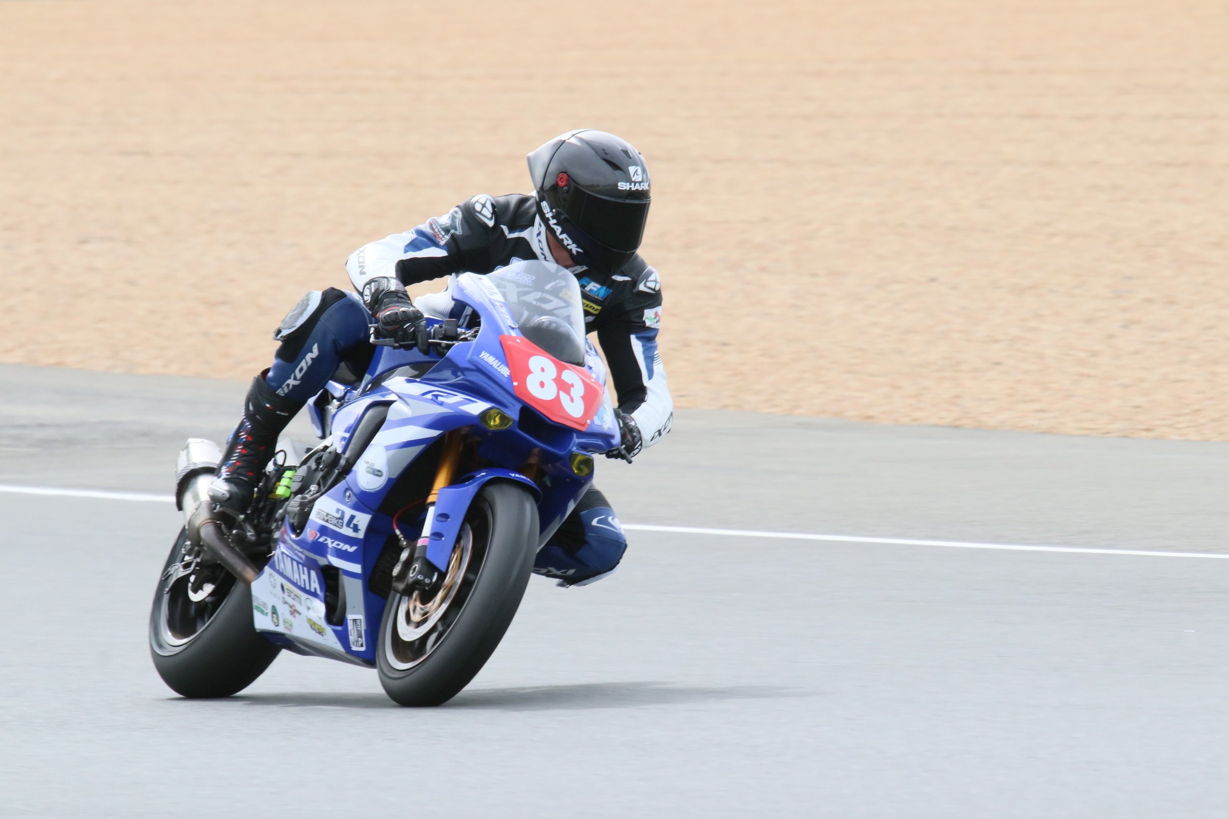 Un pilote de moto en pleine course, portant un casque noir et un équipement de protection, roulant sur une moto Yamaha bleue avec le numéro 83, sur une route de course.