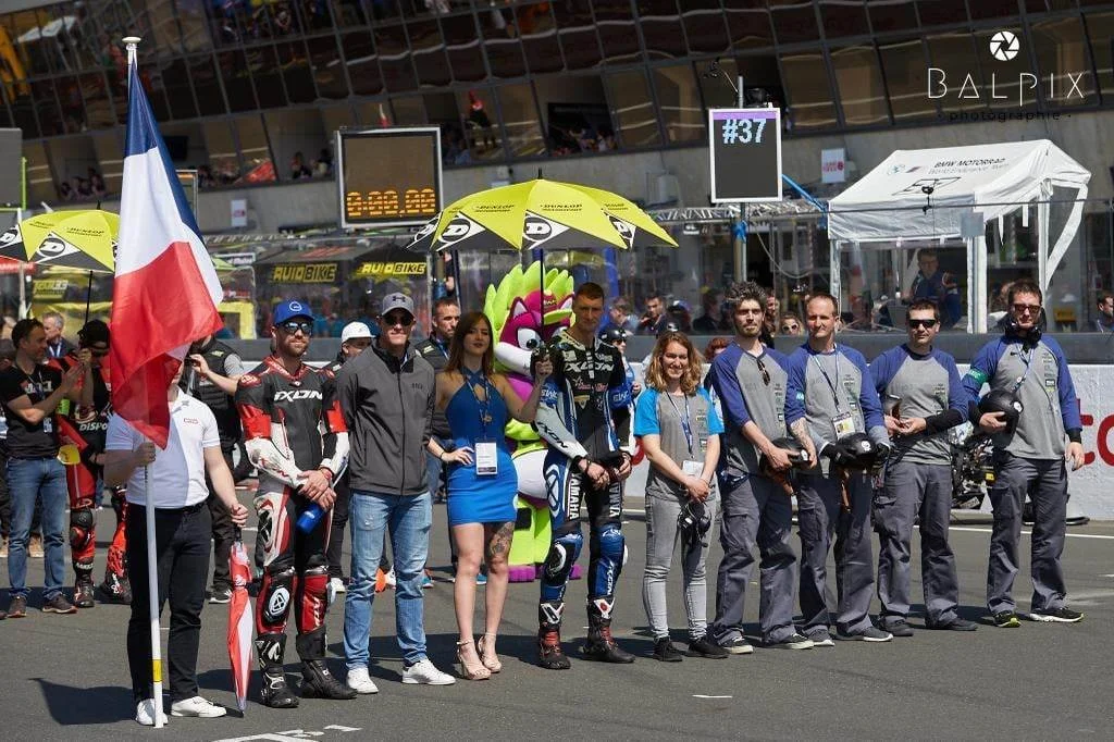 Groupe de personnes, certains en combinaisons de course, se tenant en ligne sur une piste de course lors d'un événement de moto, avec des parapluies et une mascotte colorée derrière. Un drapeau français est brandi par une personne à gauche.